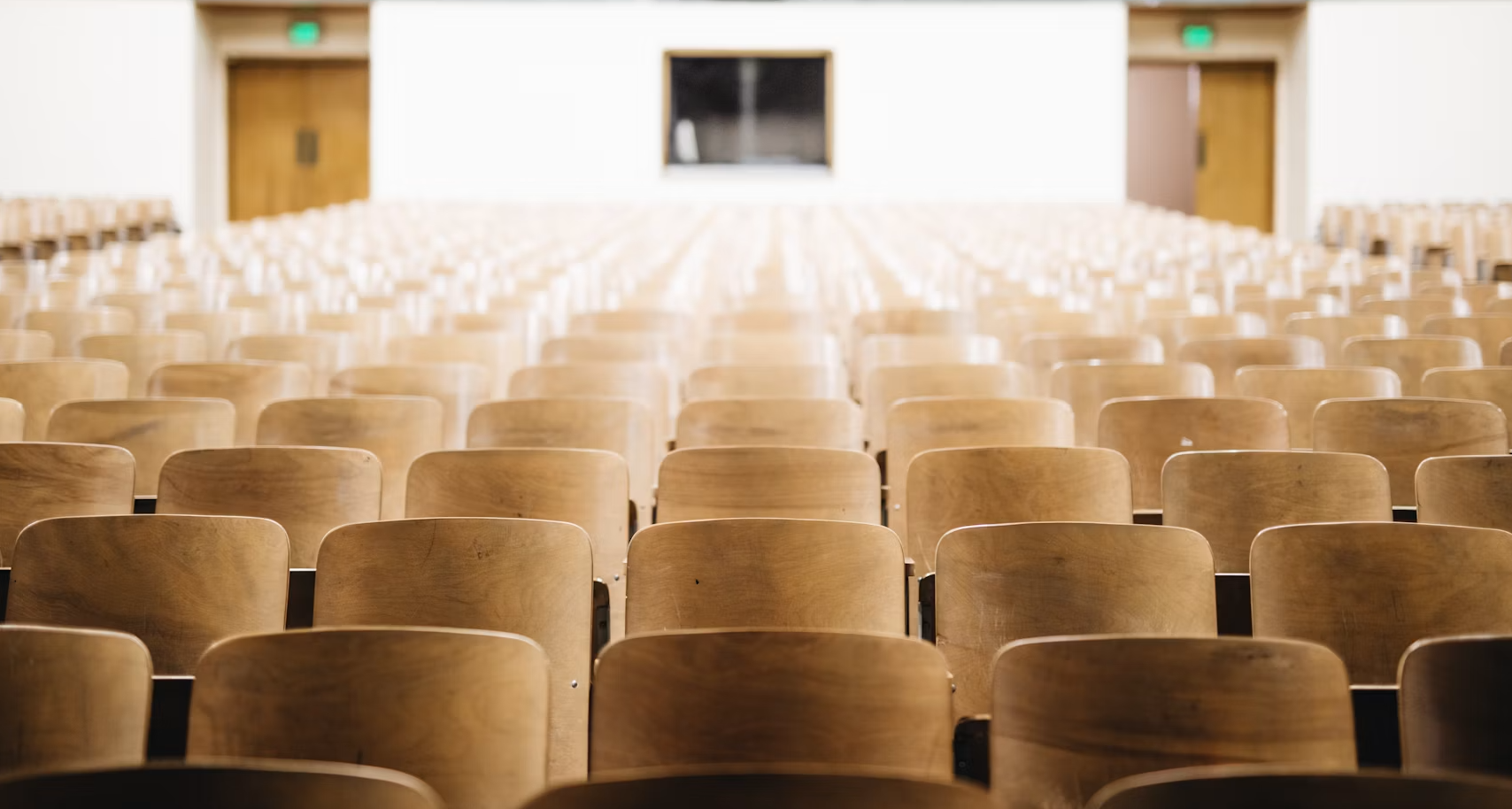 Empty wooden chairs arranged in rows in an auditorium or lecture hall with a large screen at the front.