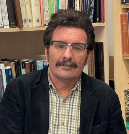 A man with dark hair, glasses, and a mustache, wearing a checkered shirt and dark blazer, sitting in front of a bookshelf filled with books.