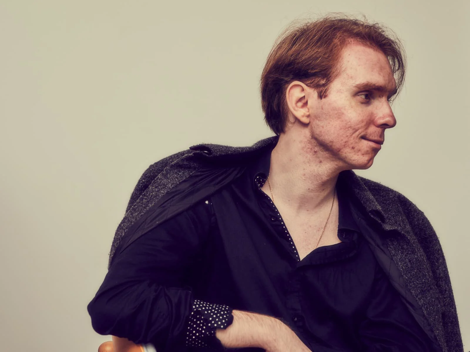 A man with short reddish hair and fair skin, wearing a dark shirt and a gray coat, is sitting against a plain, off-white background. He is looking to his right with a slight smile.