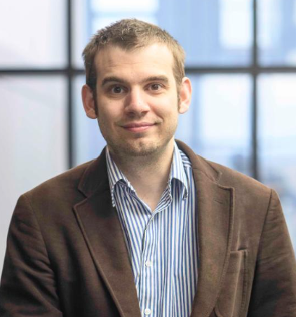 Professional man in a brown blazer and striped shirt standing in front of a windowed office background.