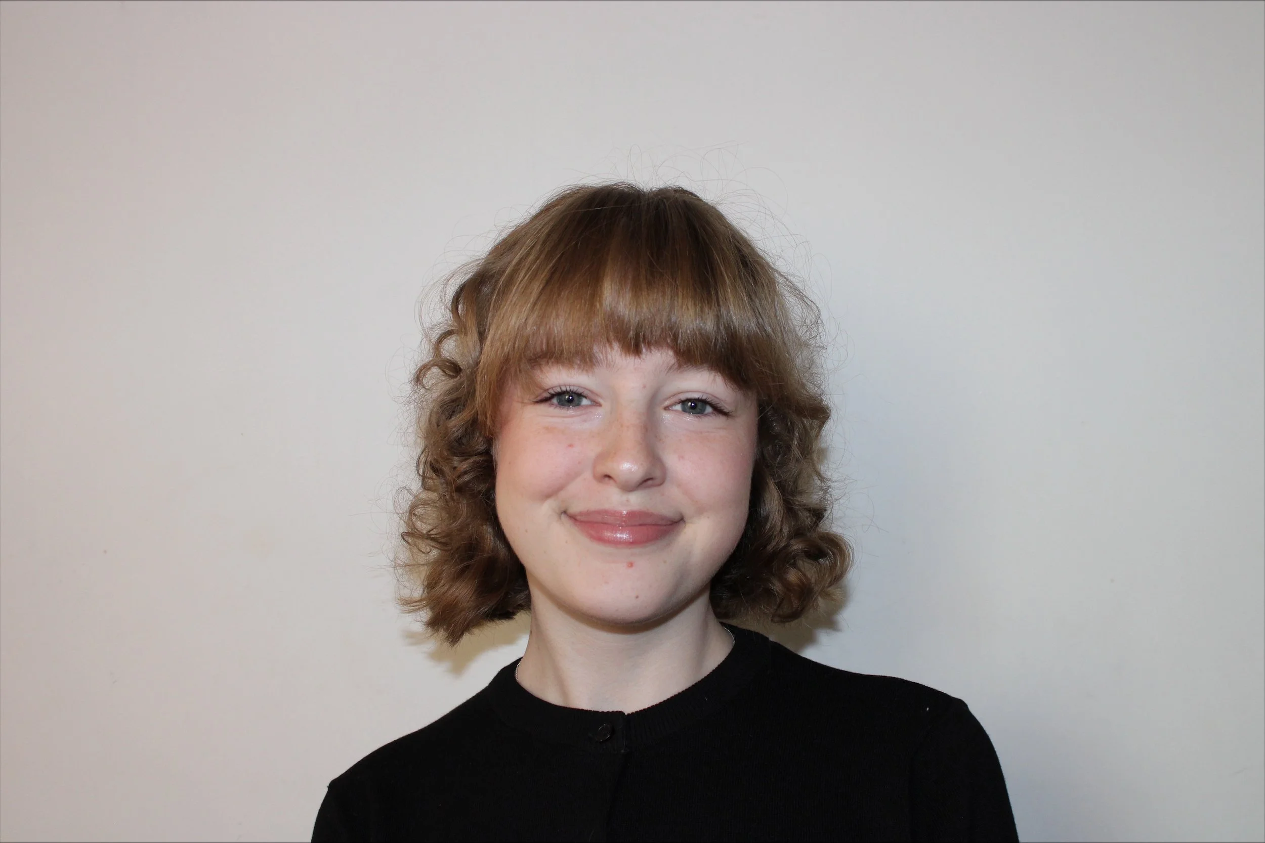 A young girl with curly light brown hair and fair skin smiles at the camera against a plain white background.