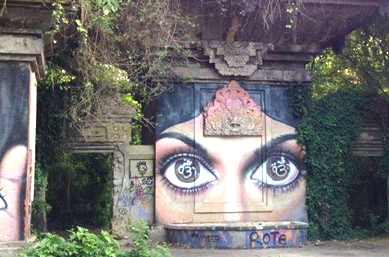 Graffiti art depicting a large pair of human eyes with a woman's face painted above the eyes on a public wall surrounded by greenery and trees.