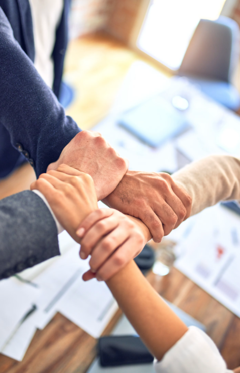 People in business attire stacking hands in teamwork gesture over a wooden table with documents and a laptop.