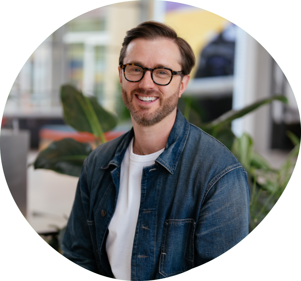 A man with glasses, brown hair, and a beard smiling in an indoor setting with plants and windows in the background.