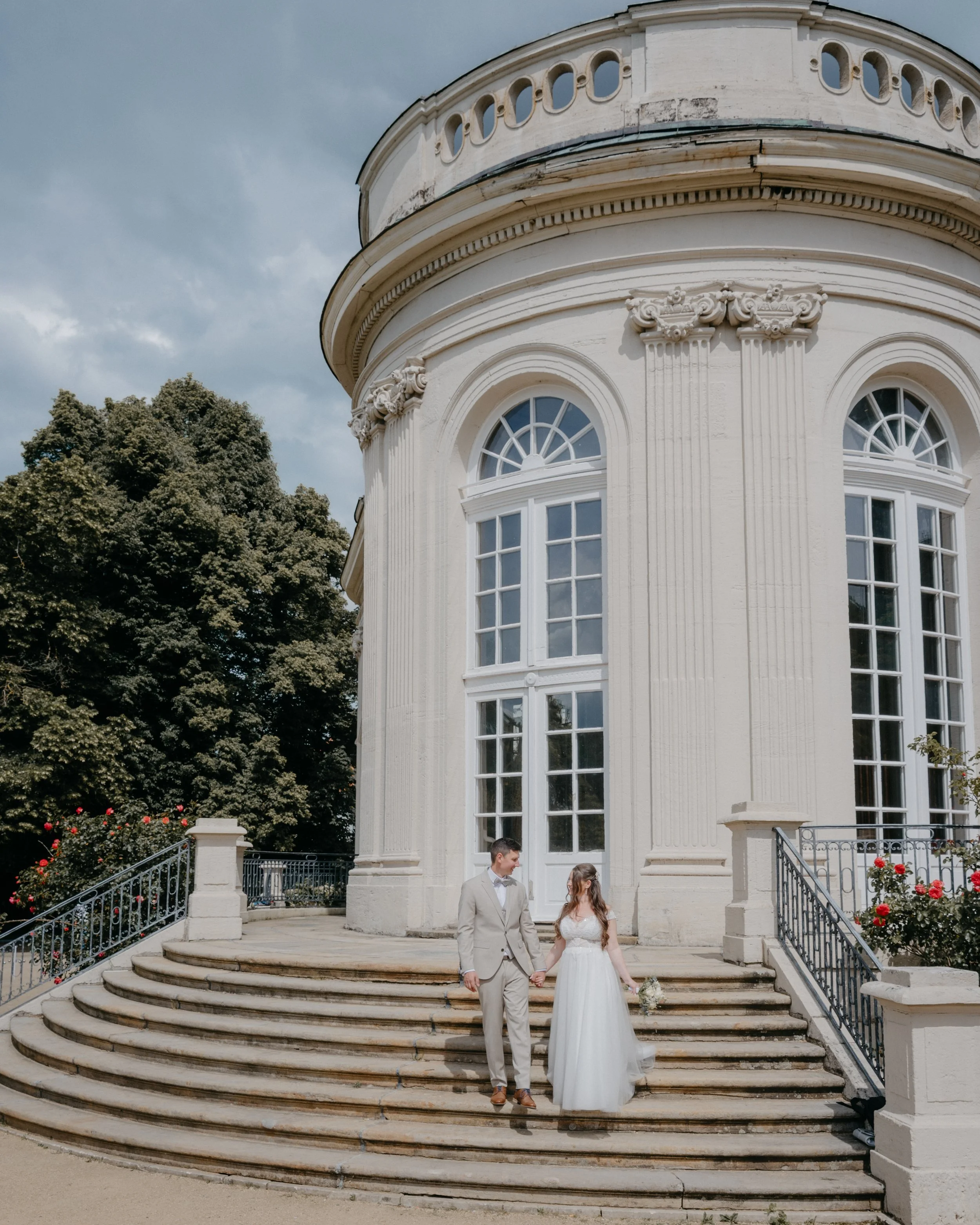 A bride and groom standing on the steps of a white, classical-style building with large windows and decorative columns, holding hands and looking at each other.