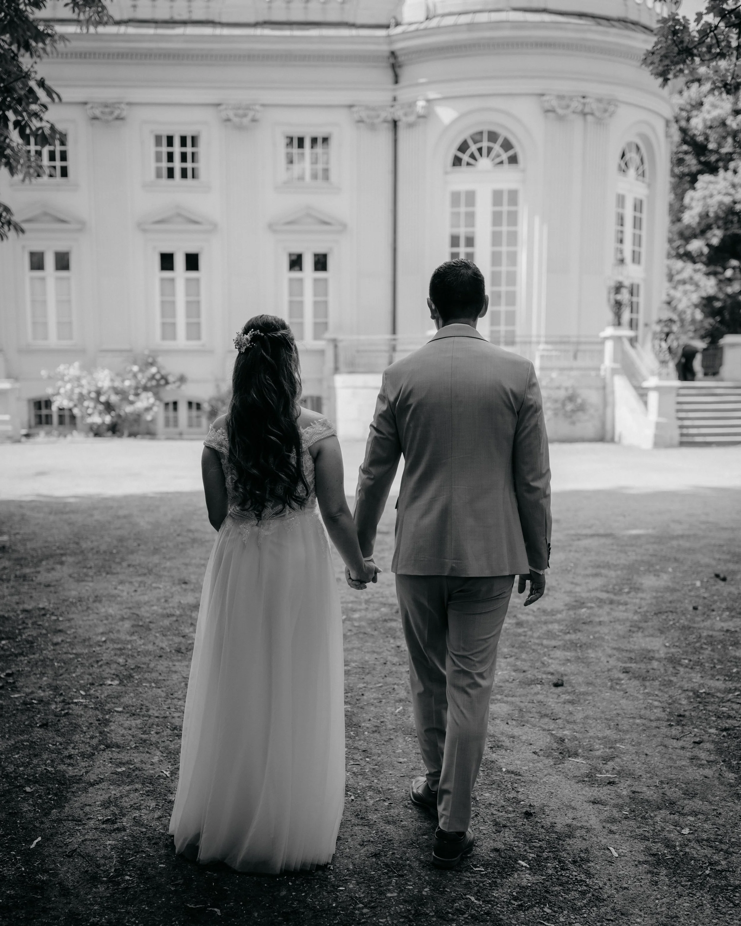 A black and white photo of a bride and groom walking hand in hand outside a large, elegant building. The bride has long, wavy hair with flowers and is wearing a wedding dress. The groom is dressed in a suit. Trees frame the scene on both sides and st