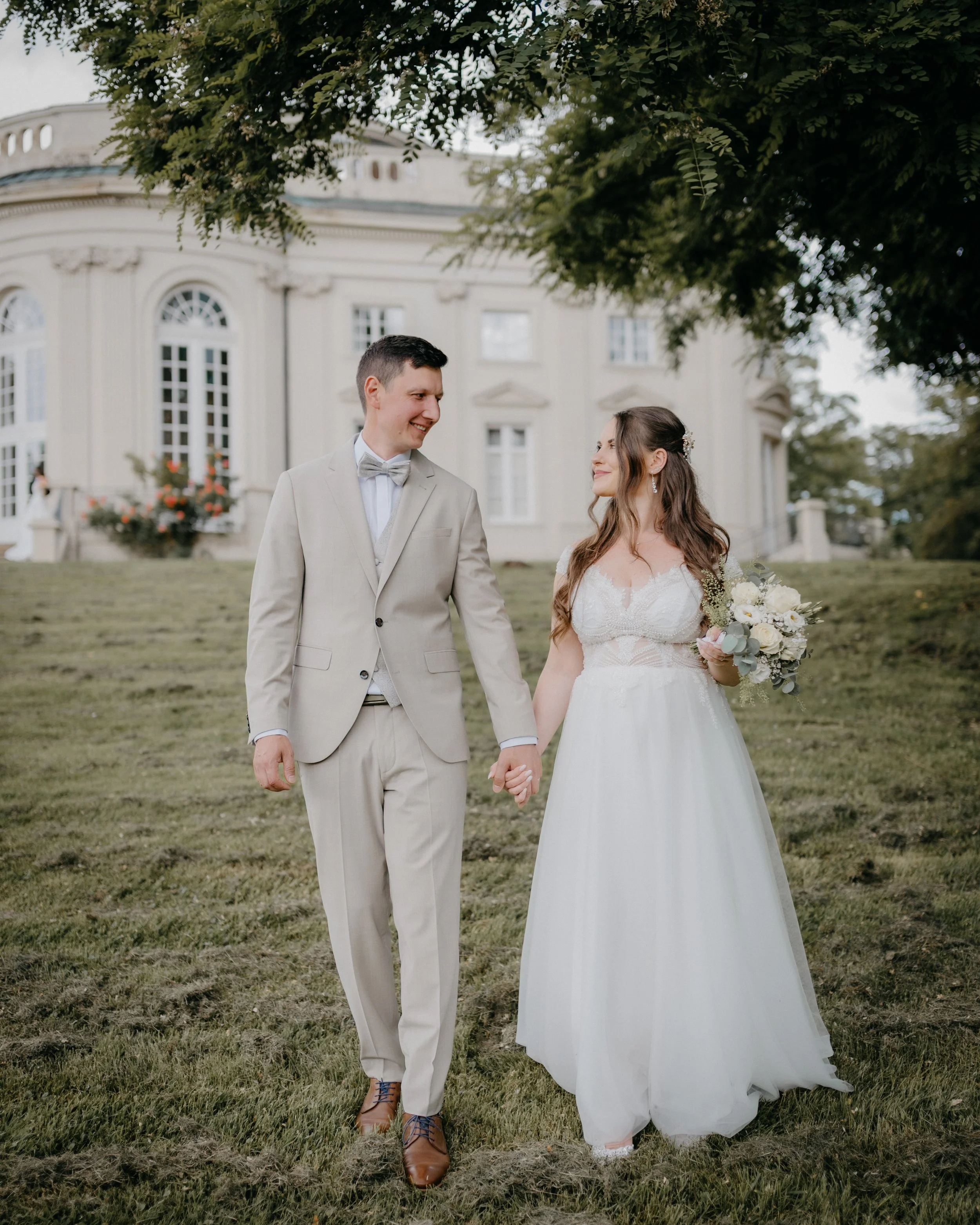 A newly married couple holding hands walking on grass in front of a large white historic building with arched windows, greenery, and a tree overhead. The groom is wearing a light gray suit, bowtie, and brown shoes. The bride is in a white wedding gow