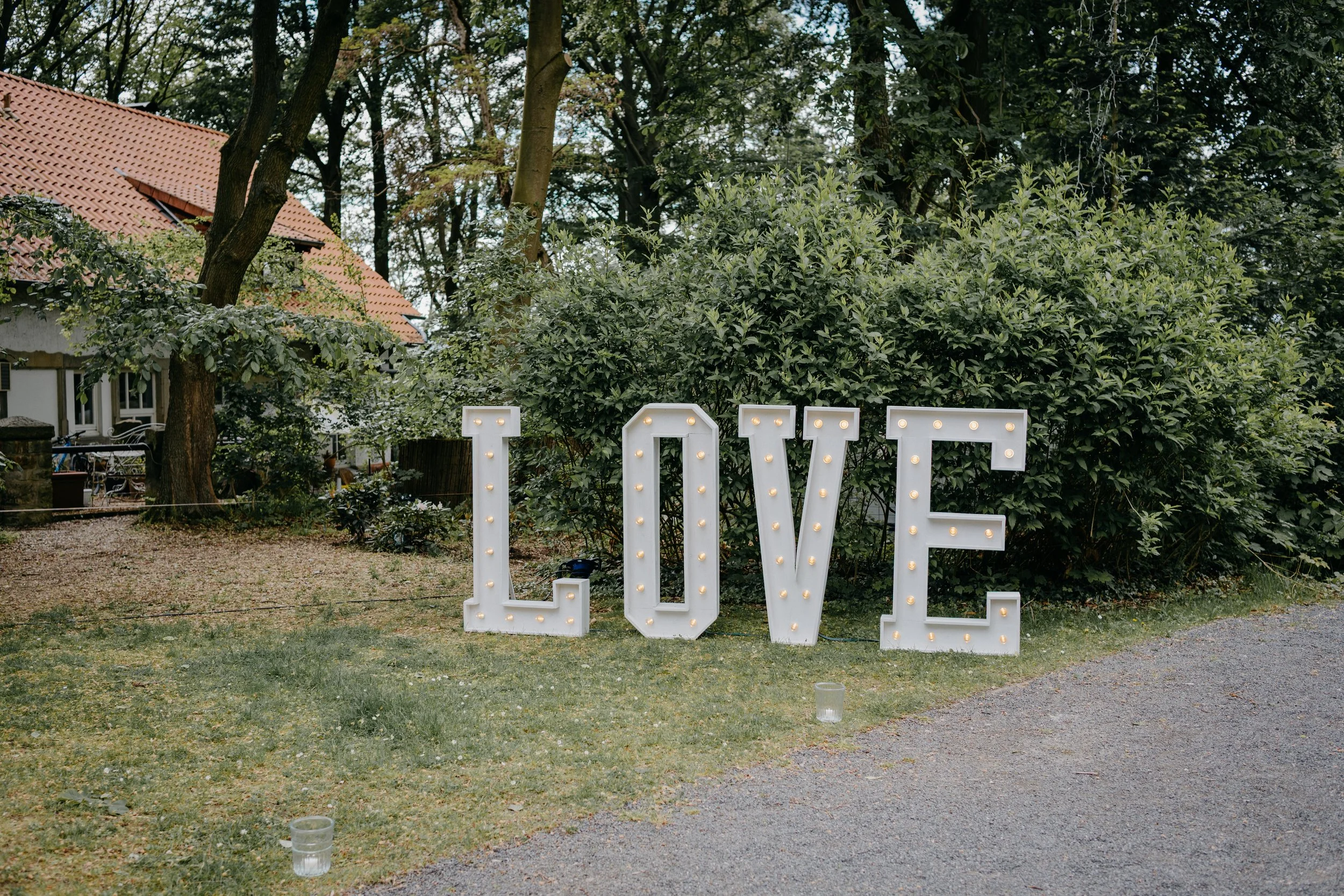 Outdoor scene with large illuminated LOVE sign on grass, trees in background, house with red tile roof visible on left, two candles on ground in front.