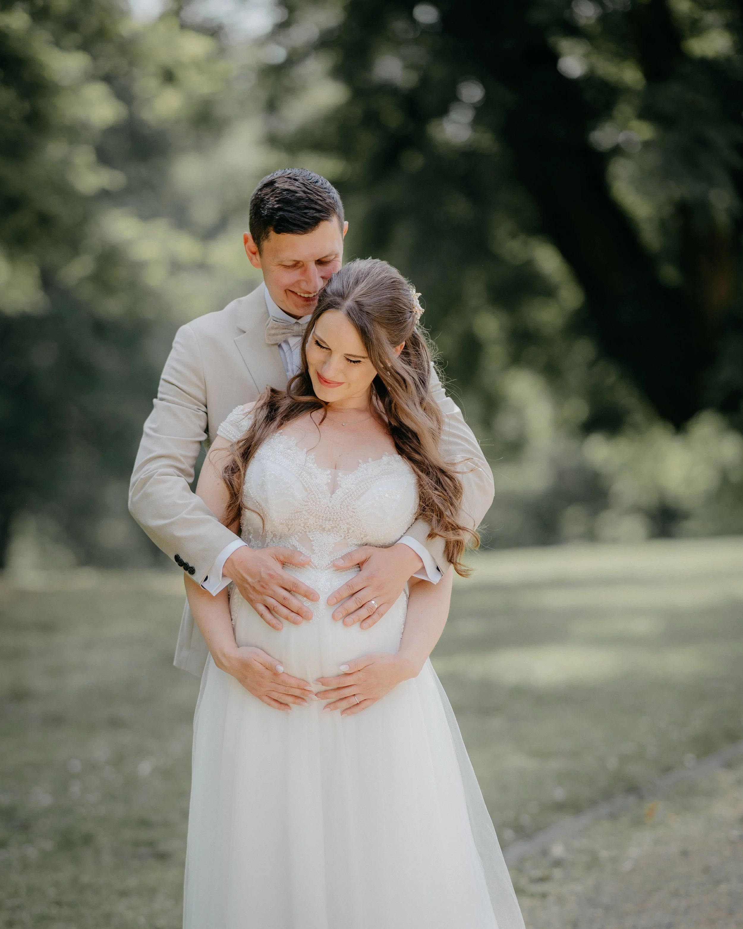 A newlywed couple standing outdoors in a park, with the groom affectionately holding the bride's belly, both smiling and looking down, surrounded by green trees.