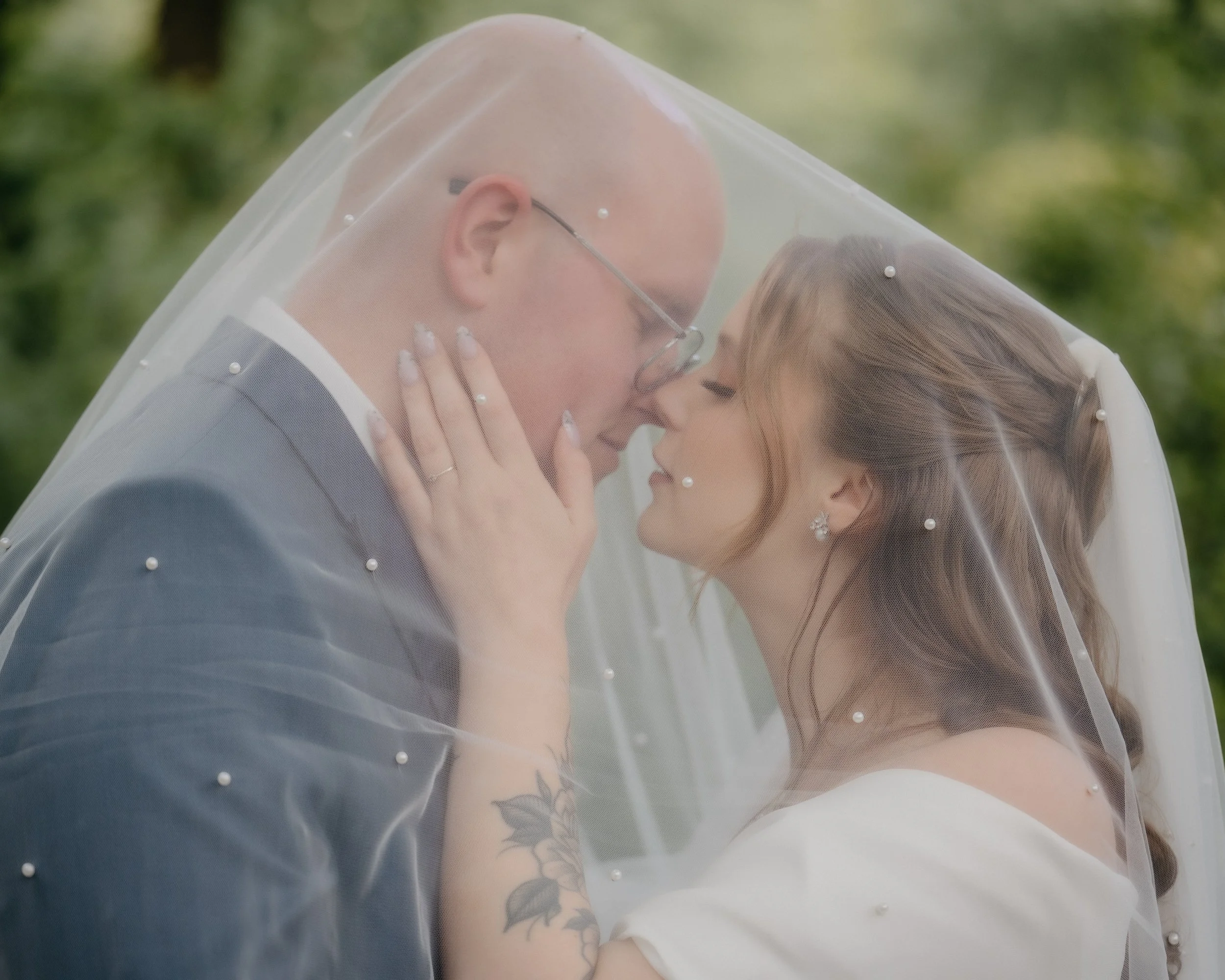 A couple sharing a kiss, covered by a sheer veil decorated with small pearls, outdoors with blurred green background.