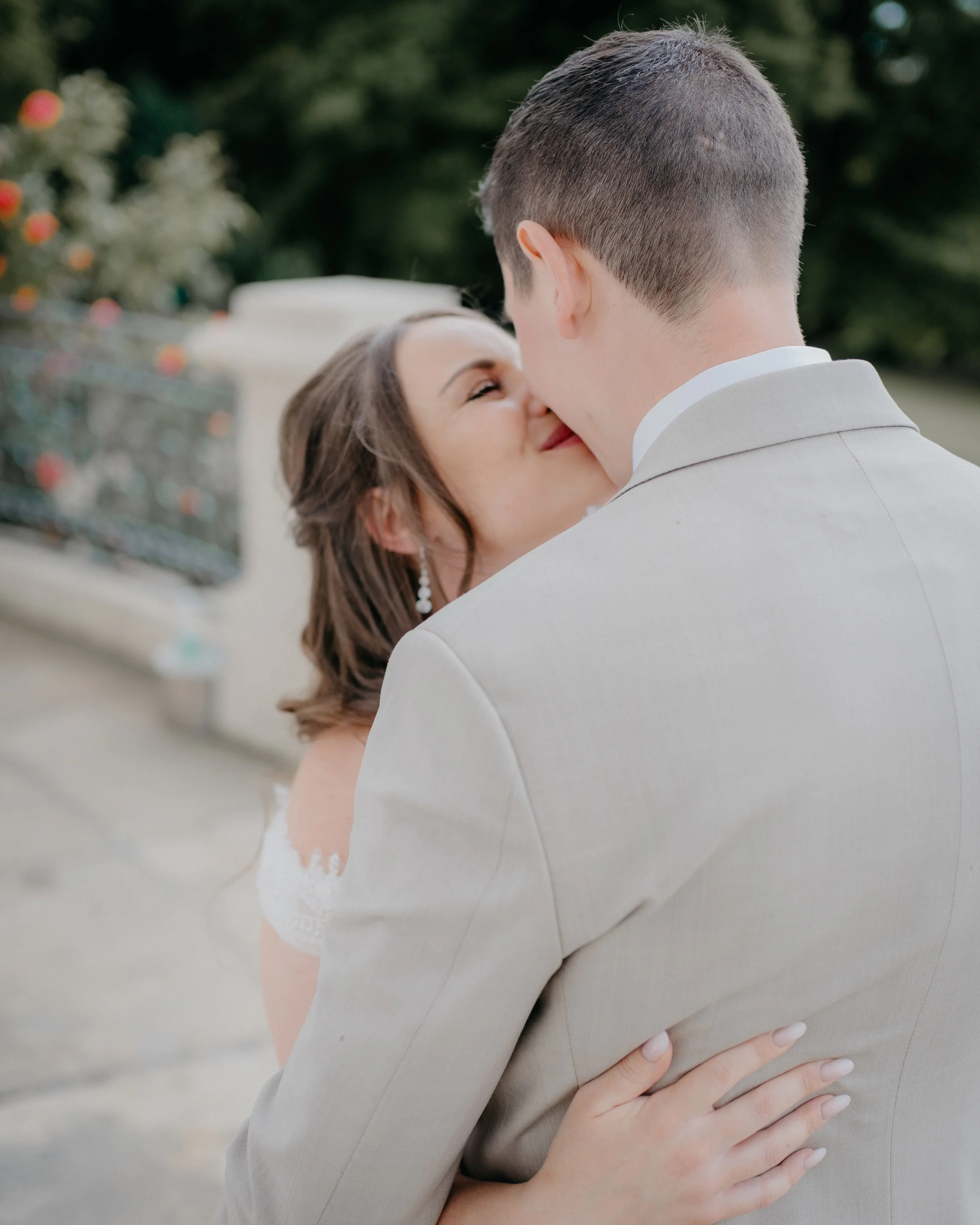 Close-up of a couple embracing outdoors, with the woman smiling and resting her head on the man's shoulder, and the man holding her close. Background of greenery and blurred flowers.
