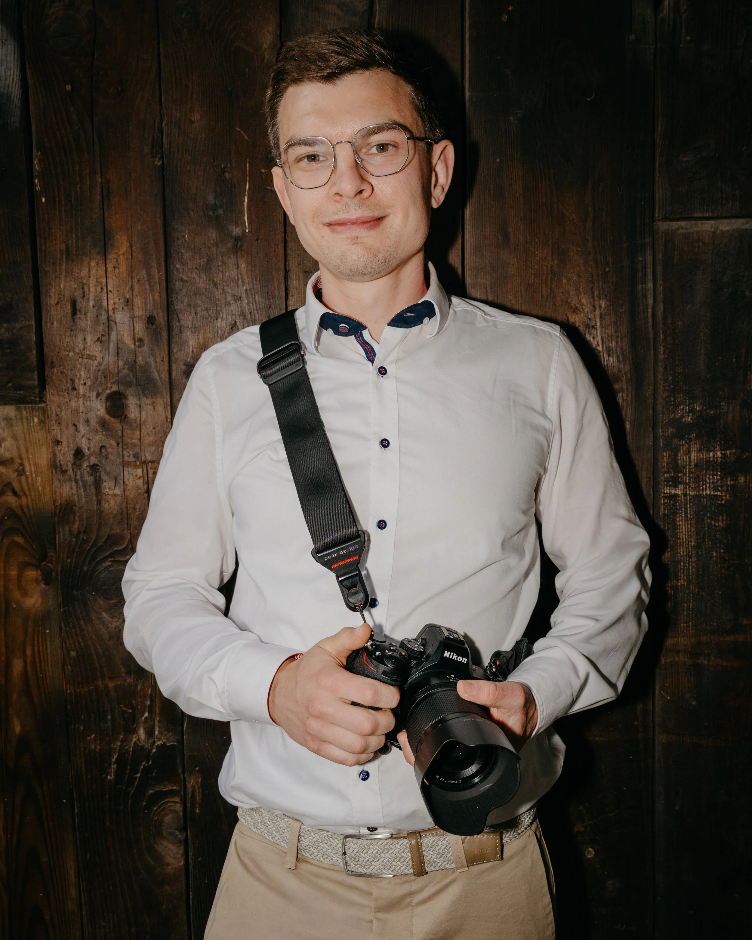 A man with glasses wearing a white shirt and beige pants, holding a Nikon camera with a large lens, standing in front of a wooden wall.