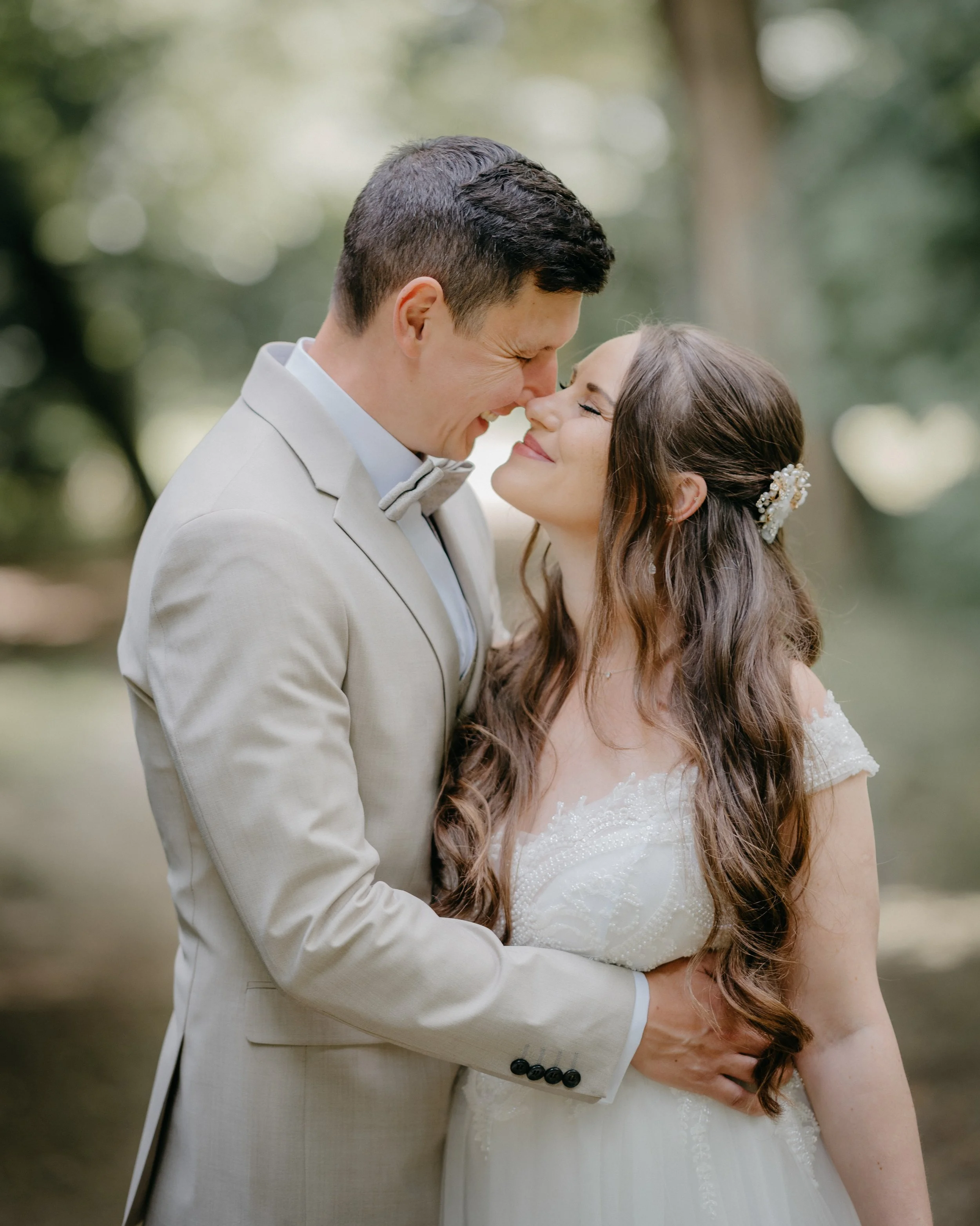 A newlywed couple smiling and touching noses outdoors with a blurred green background.