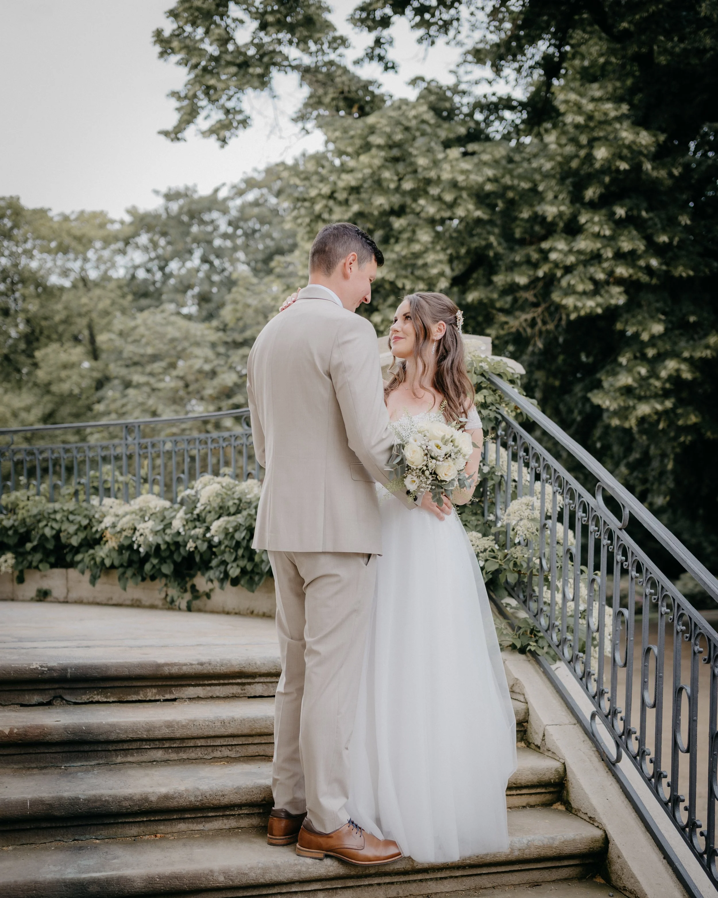 A bride and groom facing each other on outdoor staircase with greenery in background. The groom is wearing a beige suit and brown shoes, and the bride is holding a bouquet and wearing a white wedding dress with floral hair accessories.