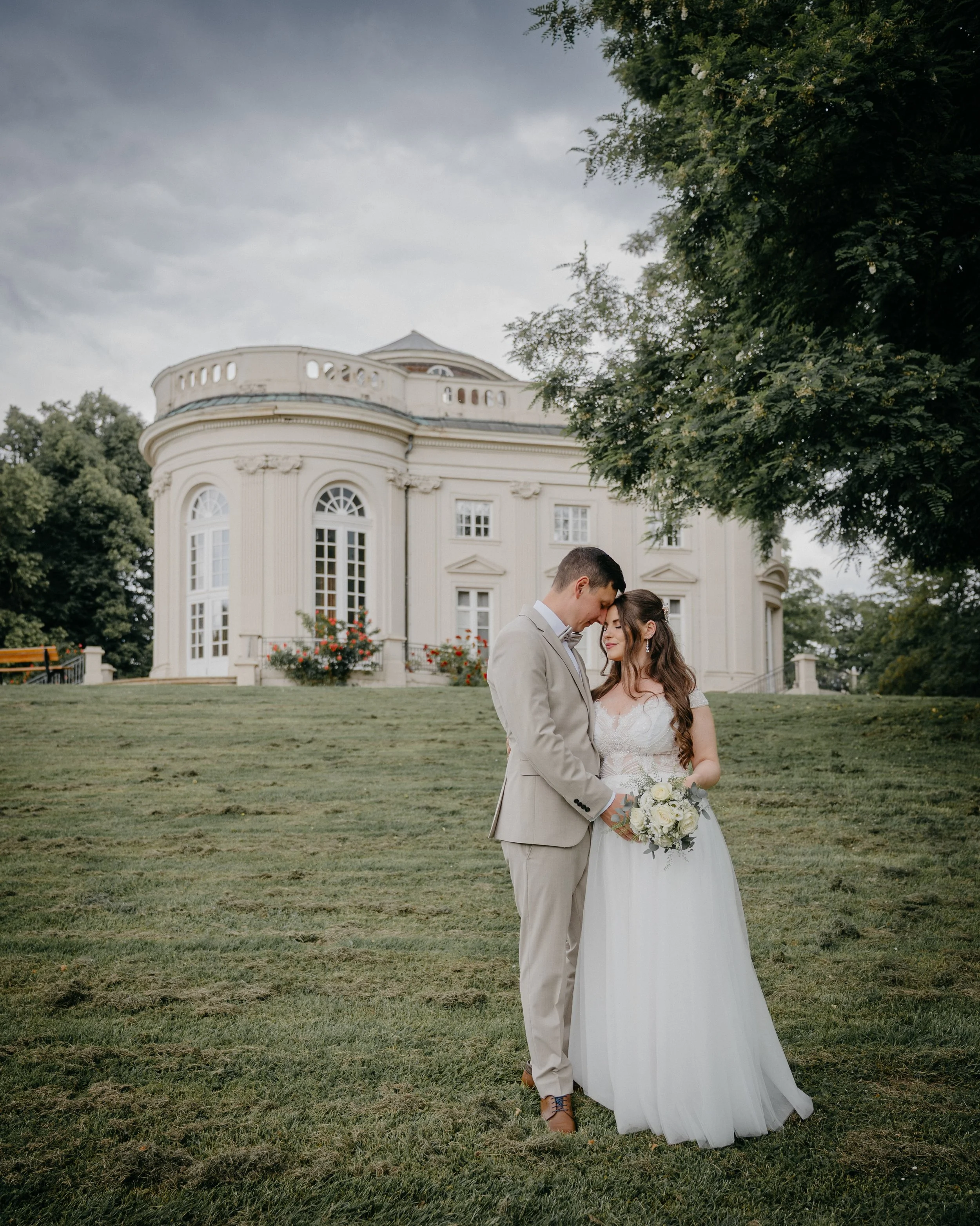 A bride and groom stand closely together outdoors on a grassy lawn, gently touching foreheads with eyes closed, in front of a large, elegant white mansion with tall windows and decorative details. The bride is wearing a white wedding gown and holding
