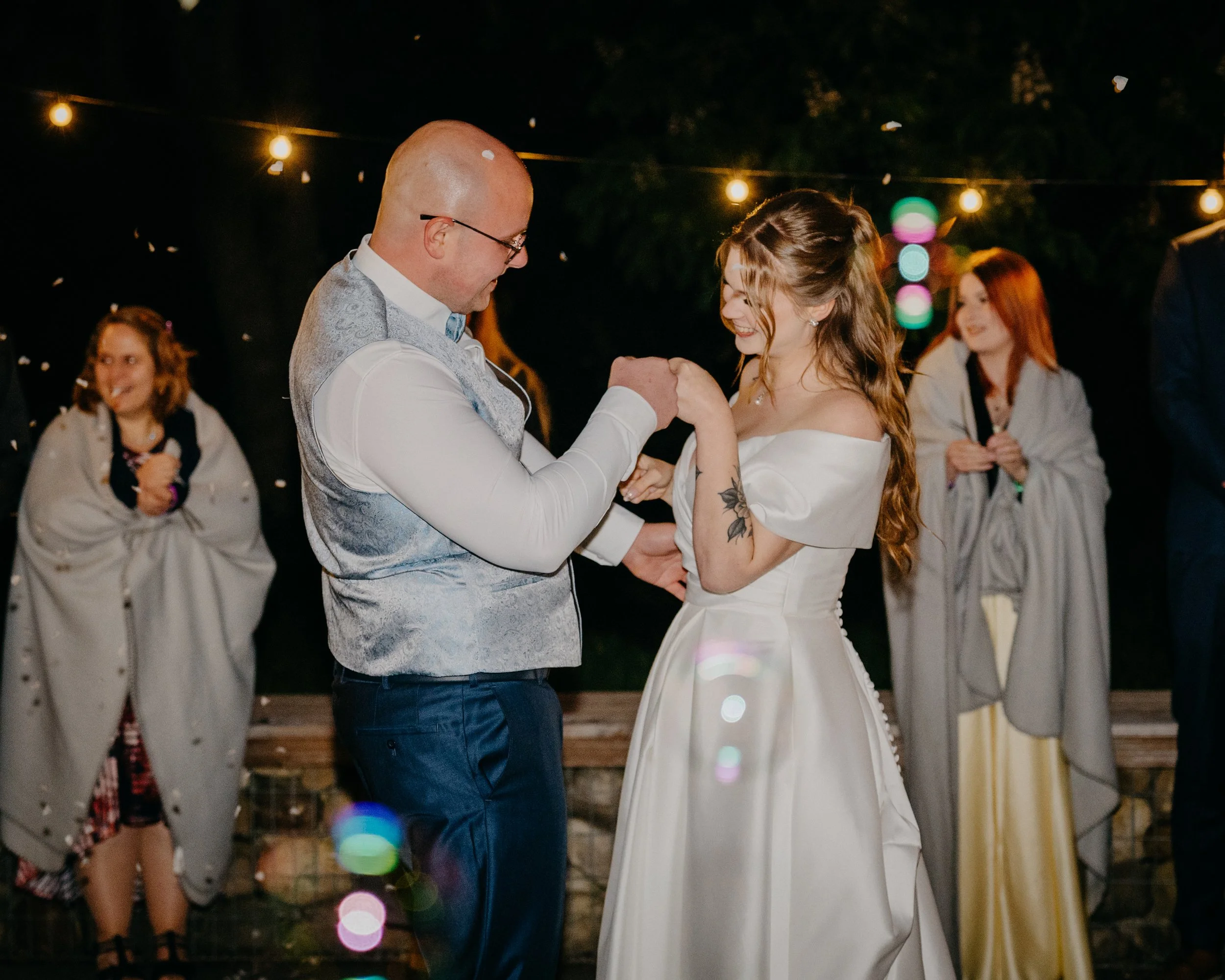 A couple dancing at an outdoor evening event with string lights overhead, surrounded by other guests, some of whom are smiling and watching.