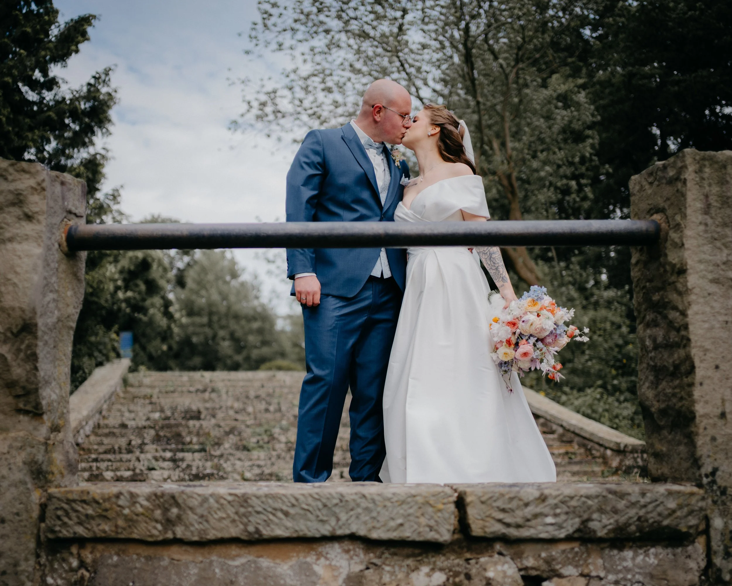 A bride and groom share a kiss outdoors on a stone staircase, with trees in the background.