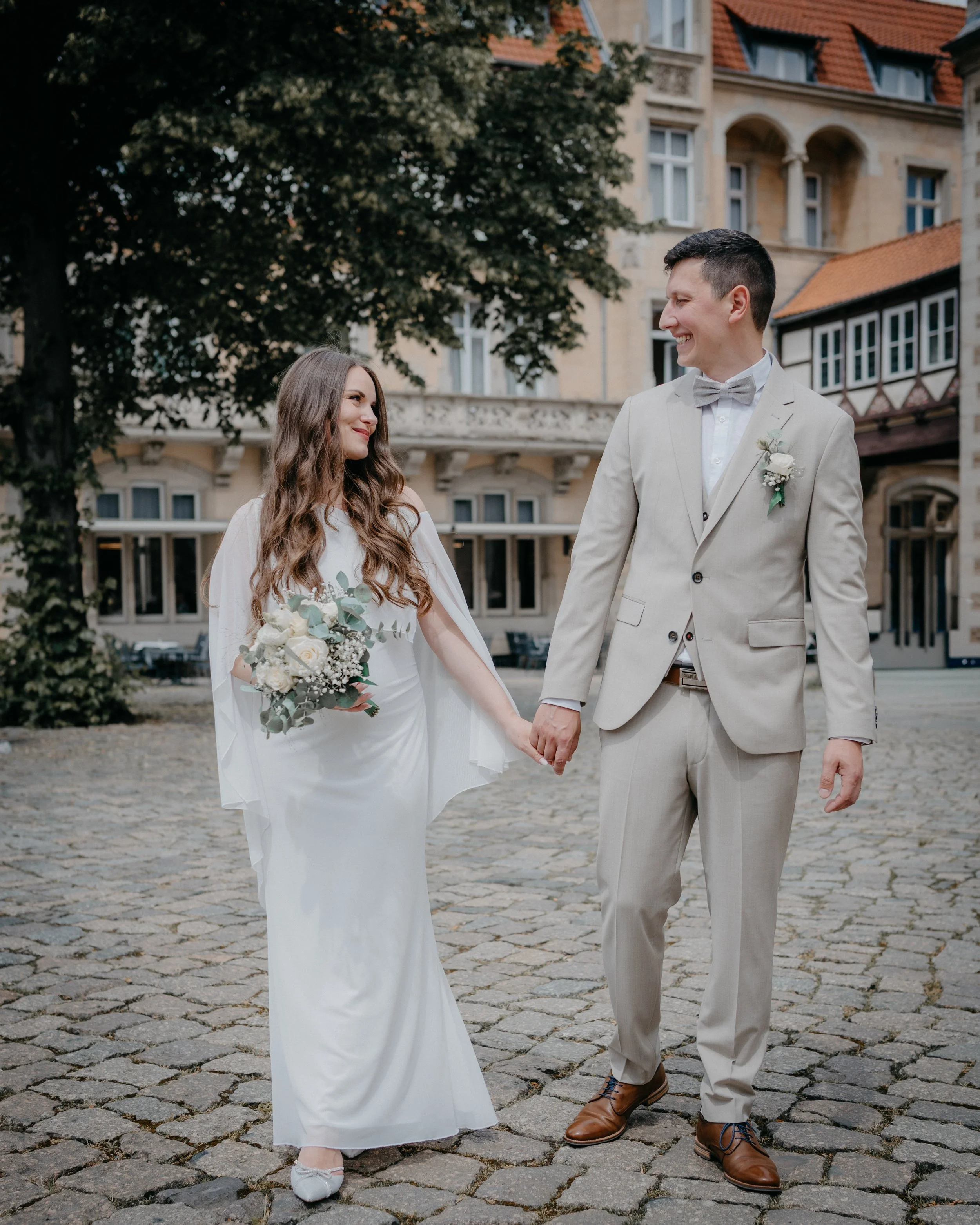 A bride and groom holding hands and smiling at each other outdoors on a cobblestone street with historic buildings in the background.
