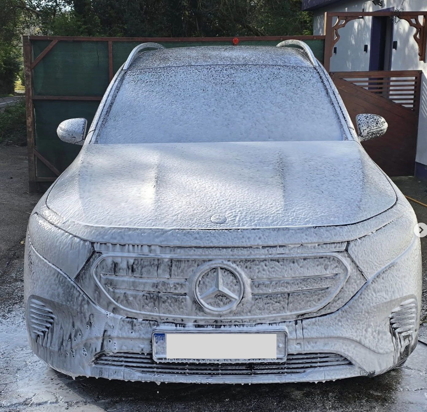 Silver Mercedes-Benz car covered in white foam soap during car wash, parked outdoors with a house and trees in the background.