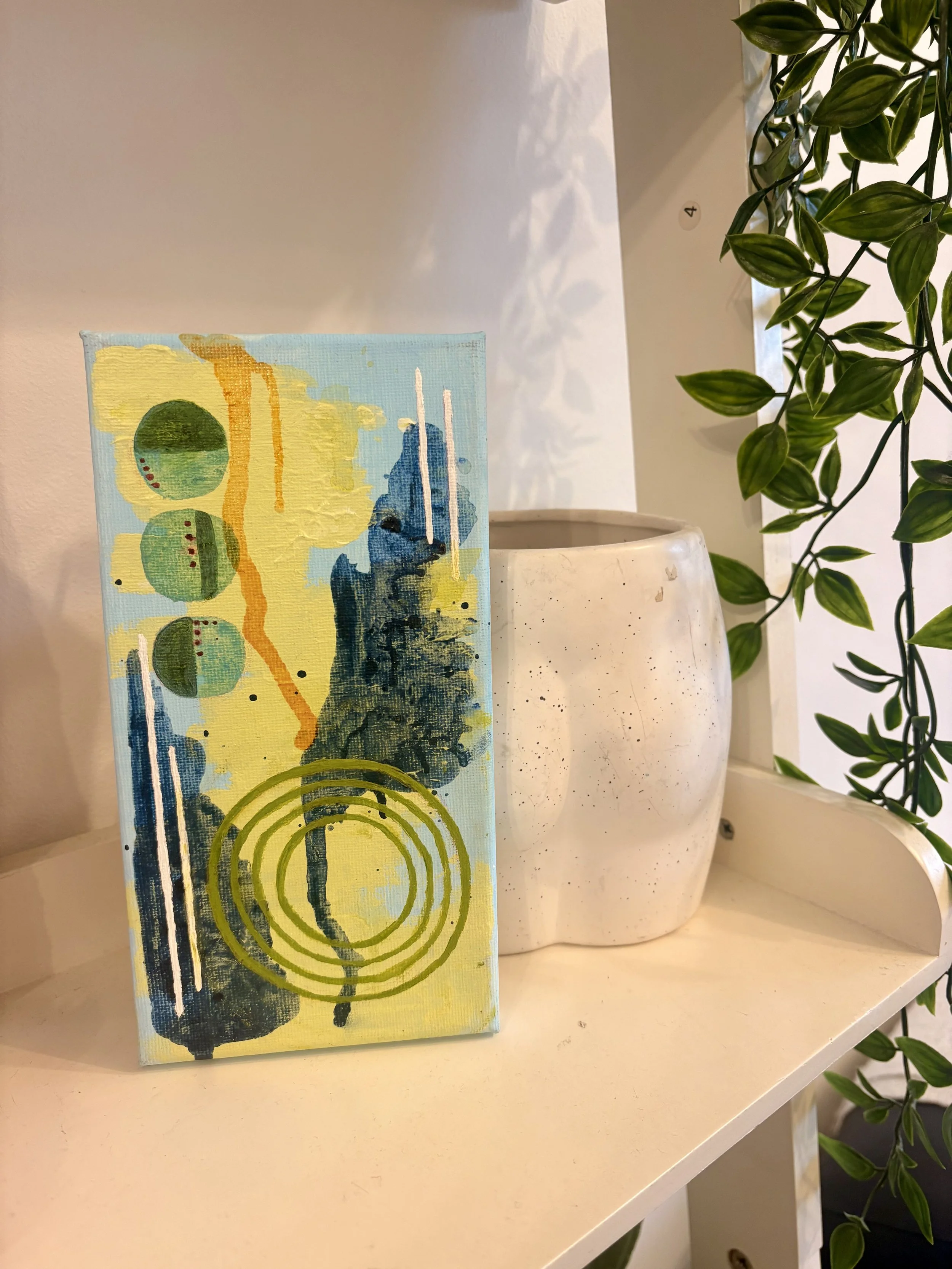 Close-up of an abstract painting on a small canvas with yellow, green, and blue colors, placed on a white shelf. To the right, there are two white vases, one taller and the other shorter, with green leafy plants hanging over the shelf.
