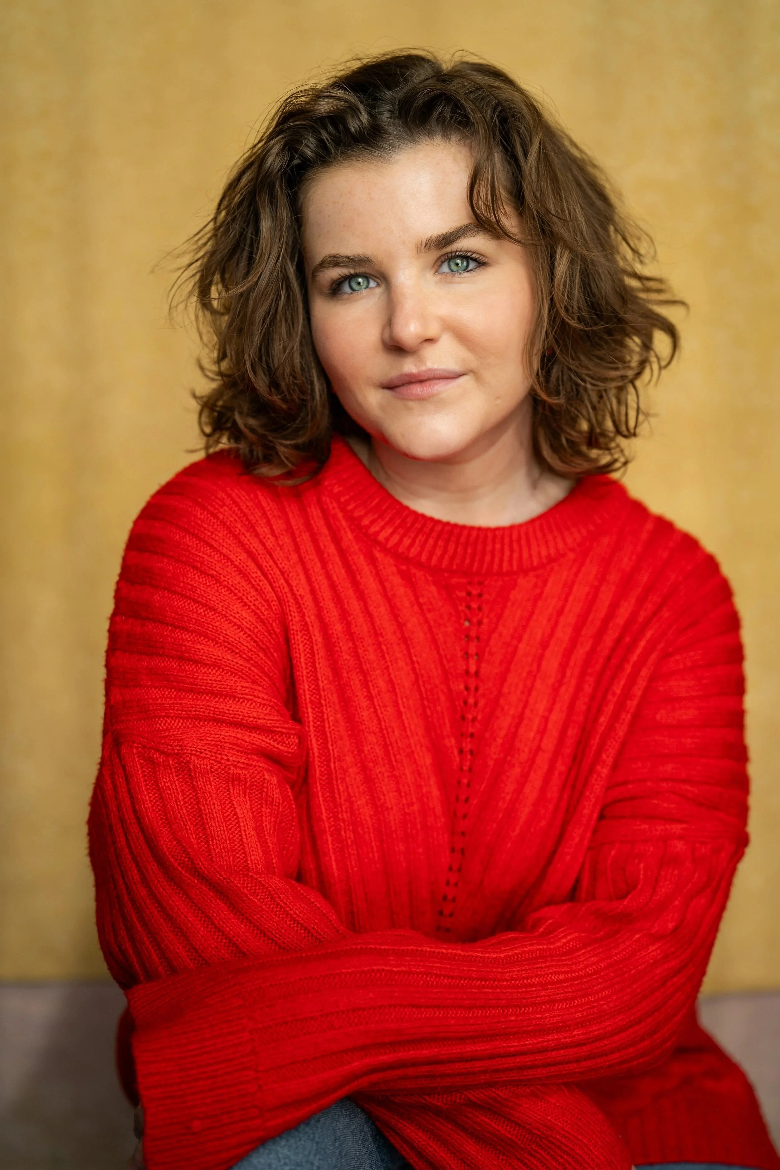 A woman with curly brown hair and blue eyes wearing a red sweater with her arms crossed in front of her, posing against a yellow background.