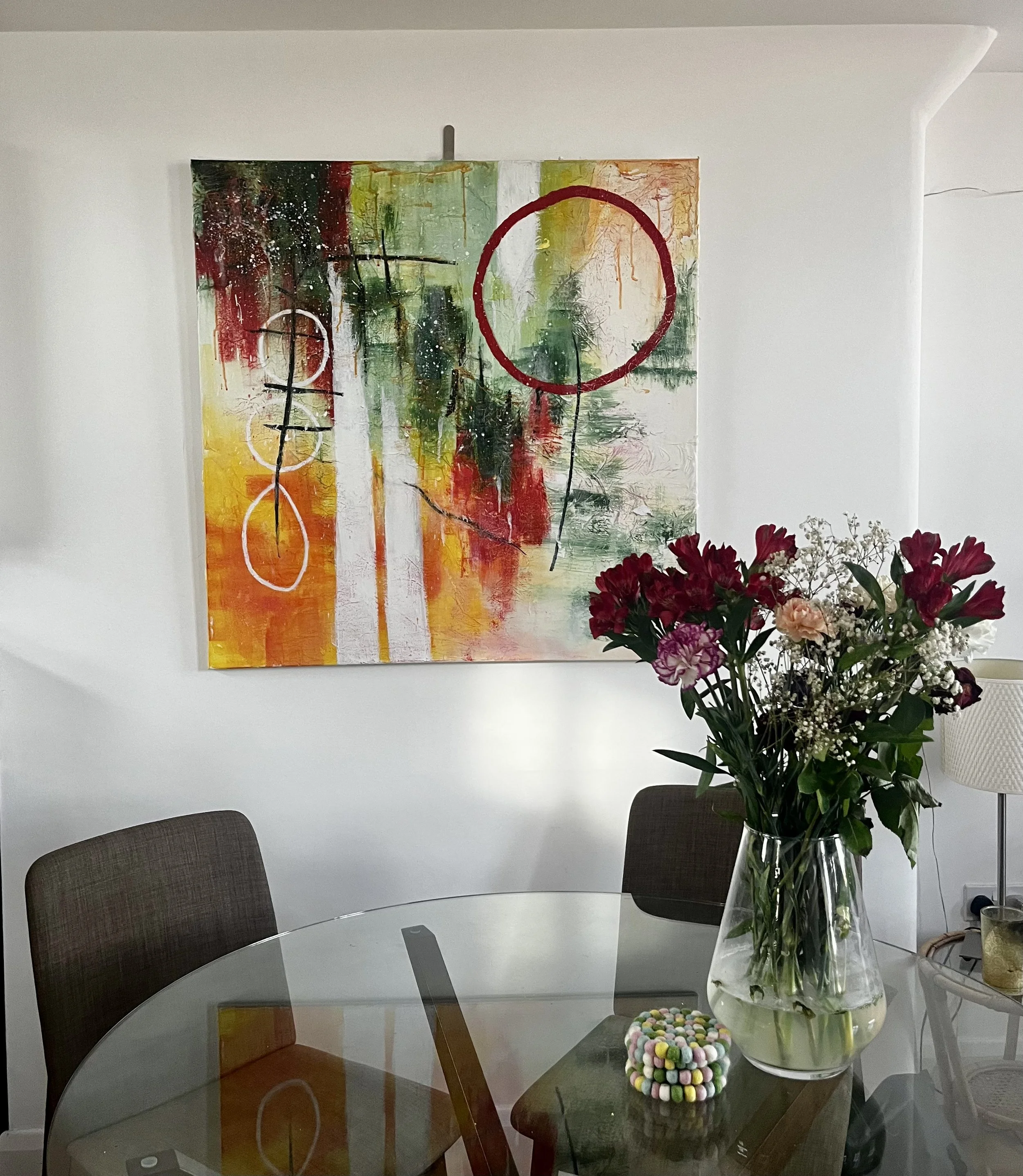A large abstract painting with vibrant colors and circles on a white wall. A glass table with a jar of colorful pom-poms and a vase of flowers, surrounded by chairs.
