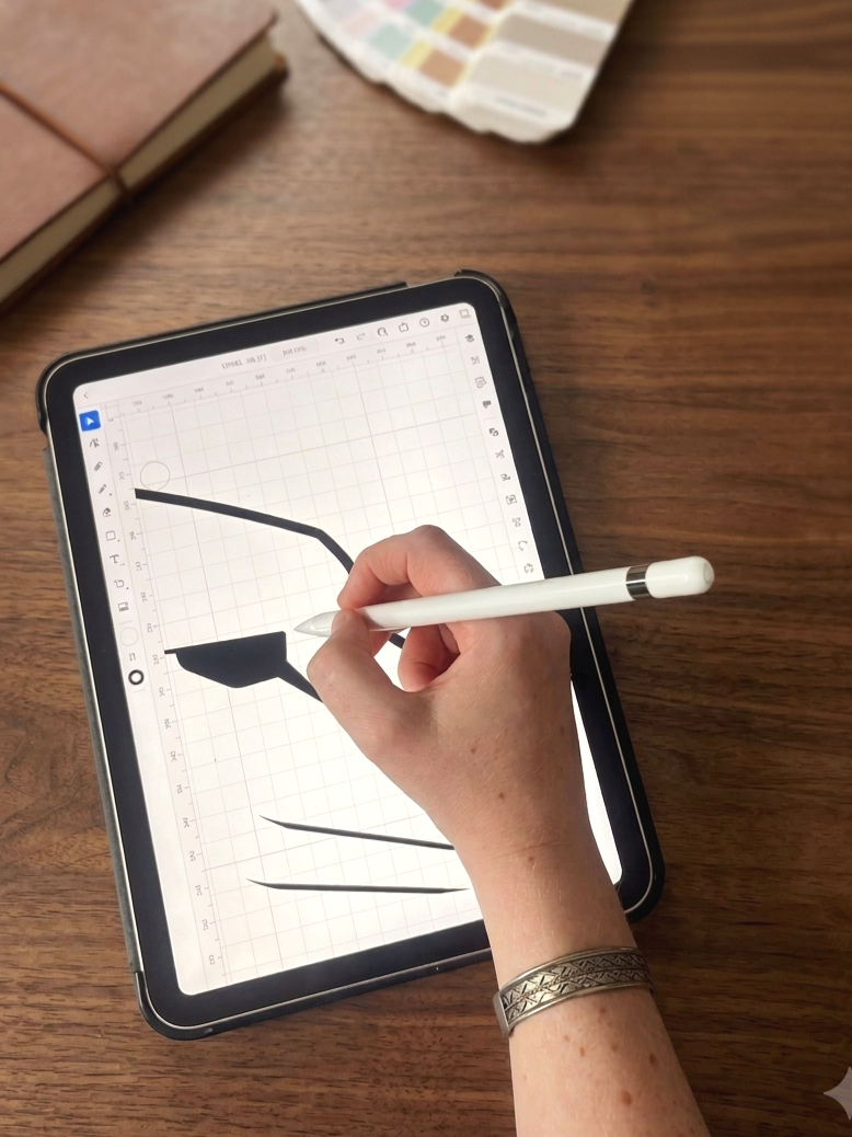 A person using a stylus to draw or write on a digital tablet that displays various black ink strokes and shapes. The tablet is on a wooden table, with a color palette card and a closed notebook nearby.