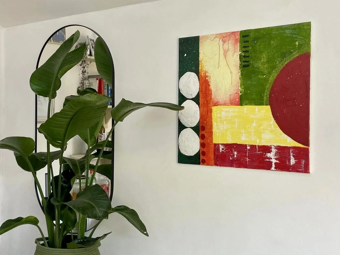 A woven basket with a large green plant beside a colorful abstract painting and a tall oval mirror on a white wall.