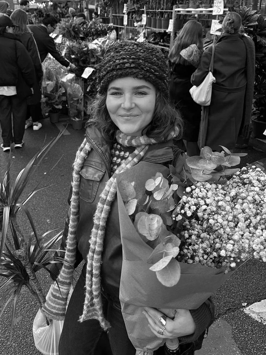 A young woman holding a bouquet of flowers at a market, smiling at the camera, surrounded by other shoppers.
