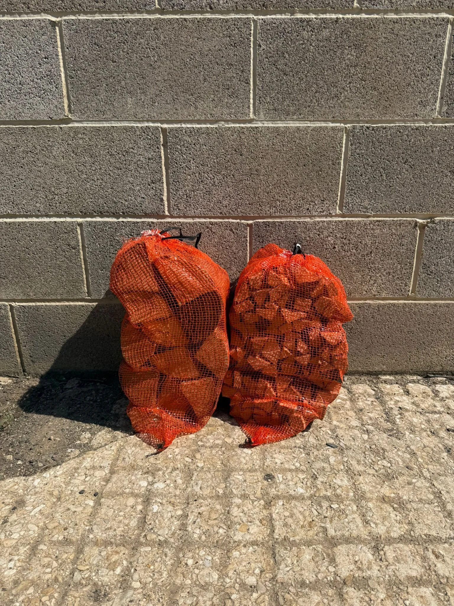 Two orange mesh bags filled with firewood and kindling, leaning against a cinder block wall.