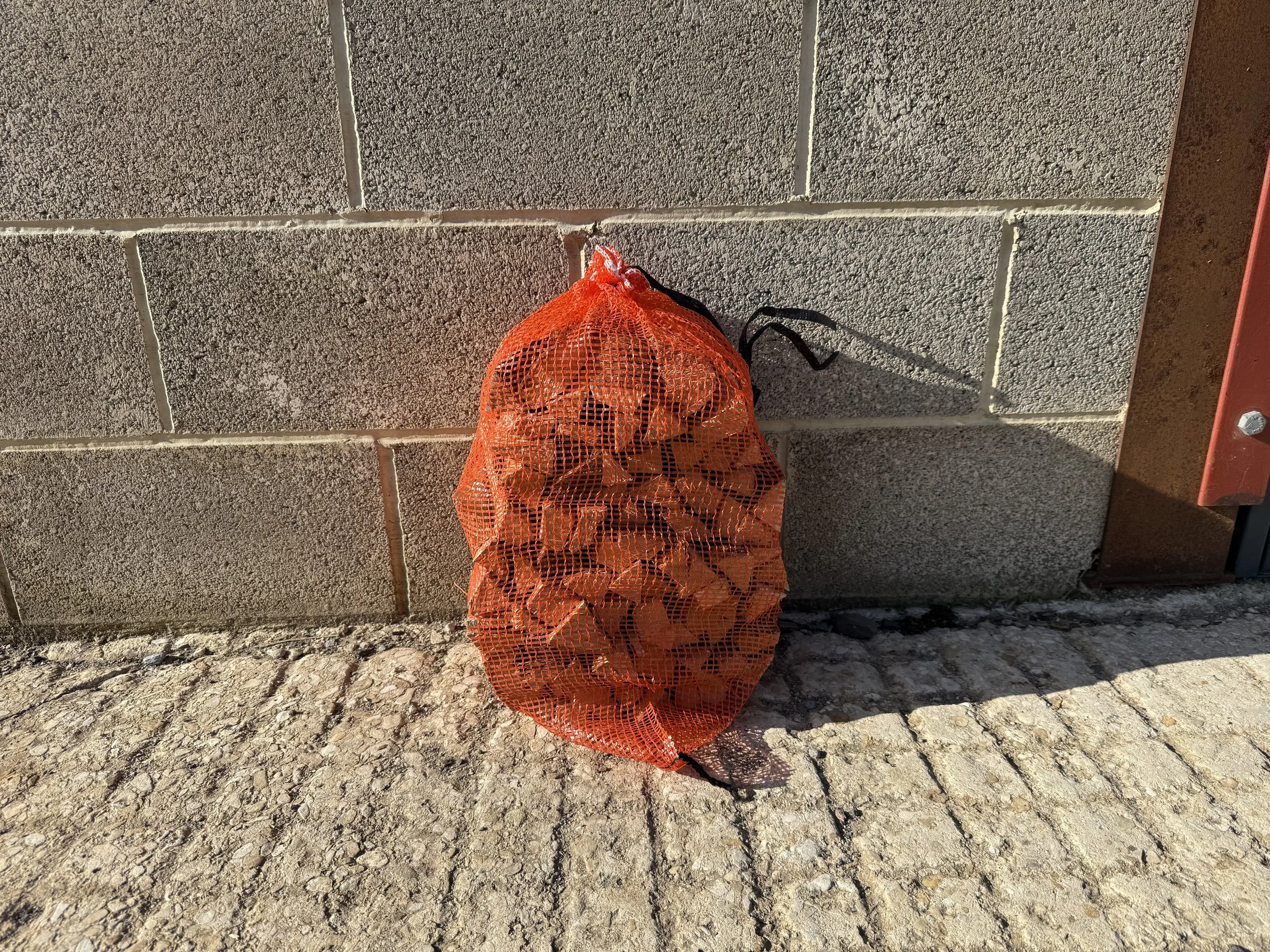 A person holding an orange mesh bag with orange pumpkins inside, standing on a rough stone pavement next to a brick wall.