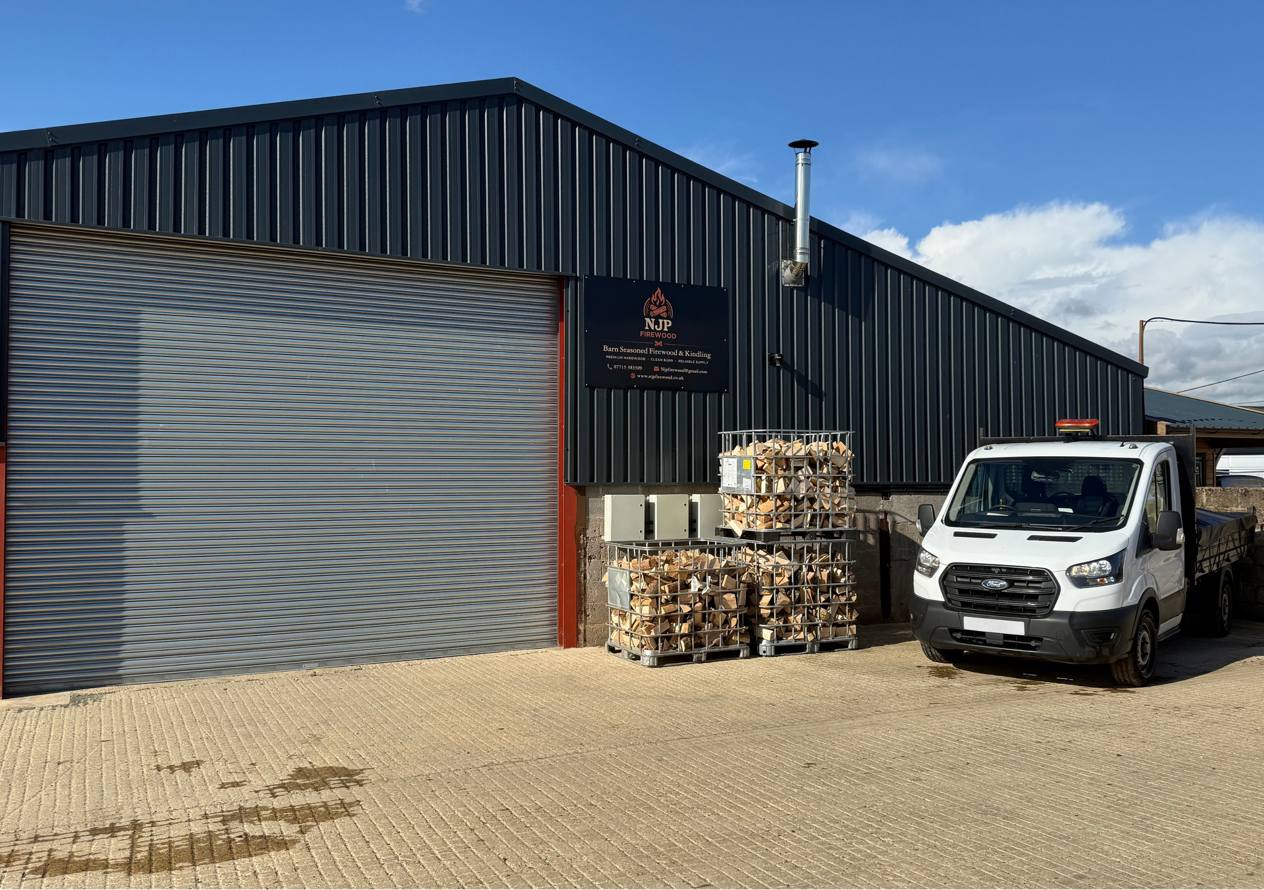 Exterior view of a commercial building with dark blue corrugated metal siding, a large closed roller shutter door, a black sign for NJP Firewood, stacks of firewood in metal racks, and a white pickup truck parked in front under partly cloudy skies.