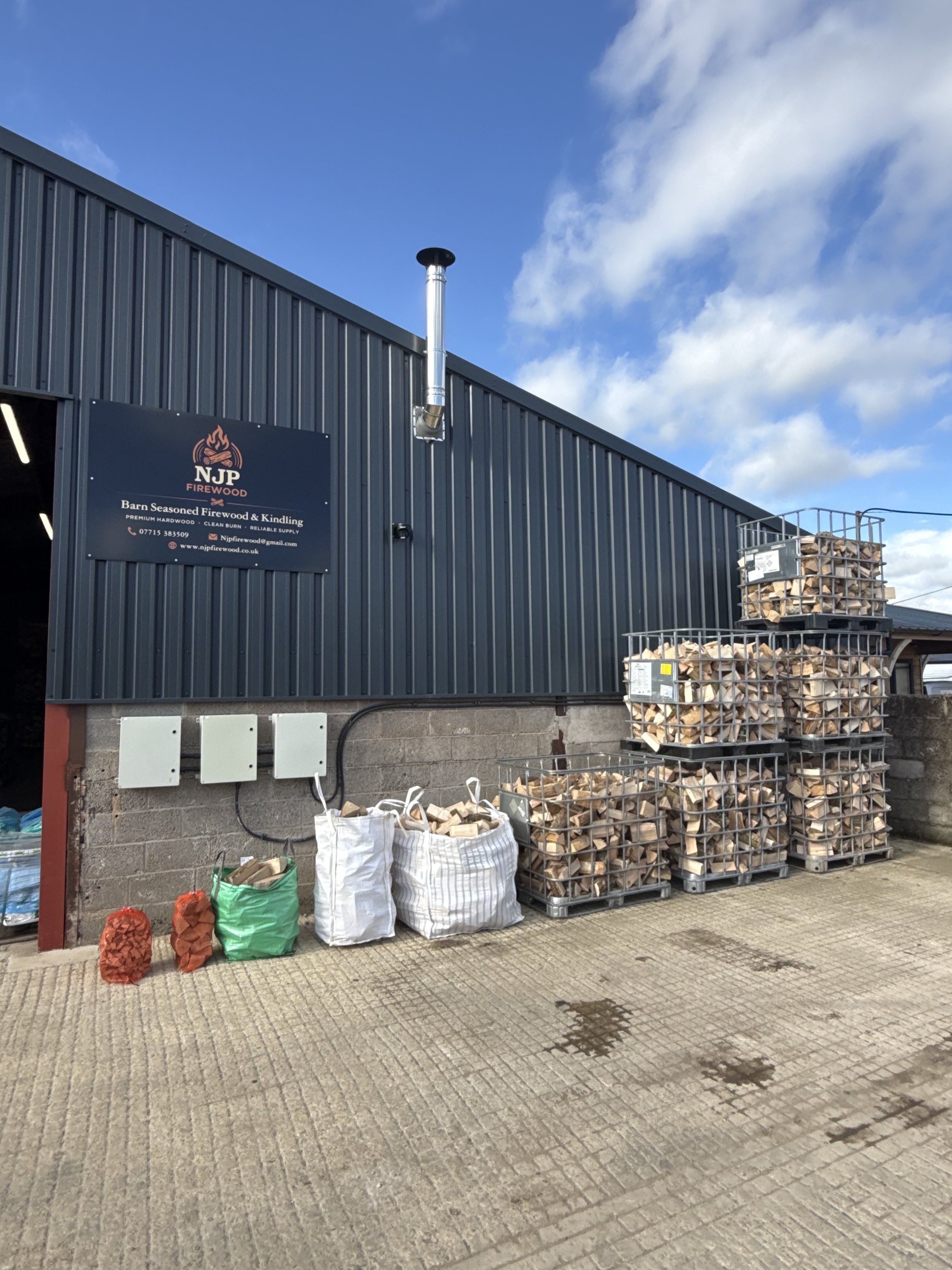 Stacked firewood in metal cages outside a building with a sign for NJP Firewood, showing bags of firewood and piles of chopped wood, against a cloudy sky.