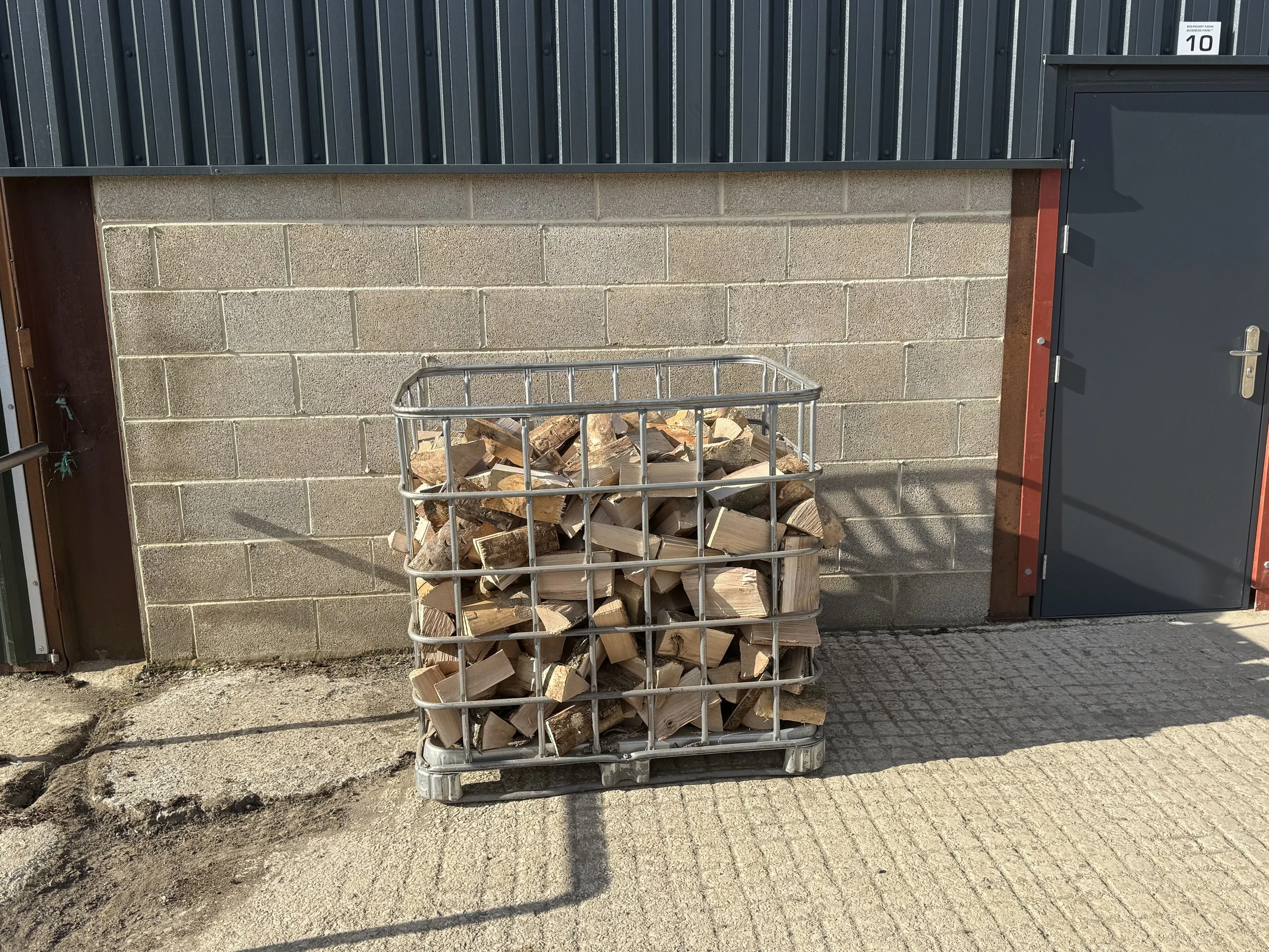 A metal basket filled with chopped firewood outside a building with a cinder block wall, a gray metal door, and black corrugated siding.