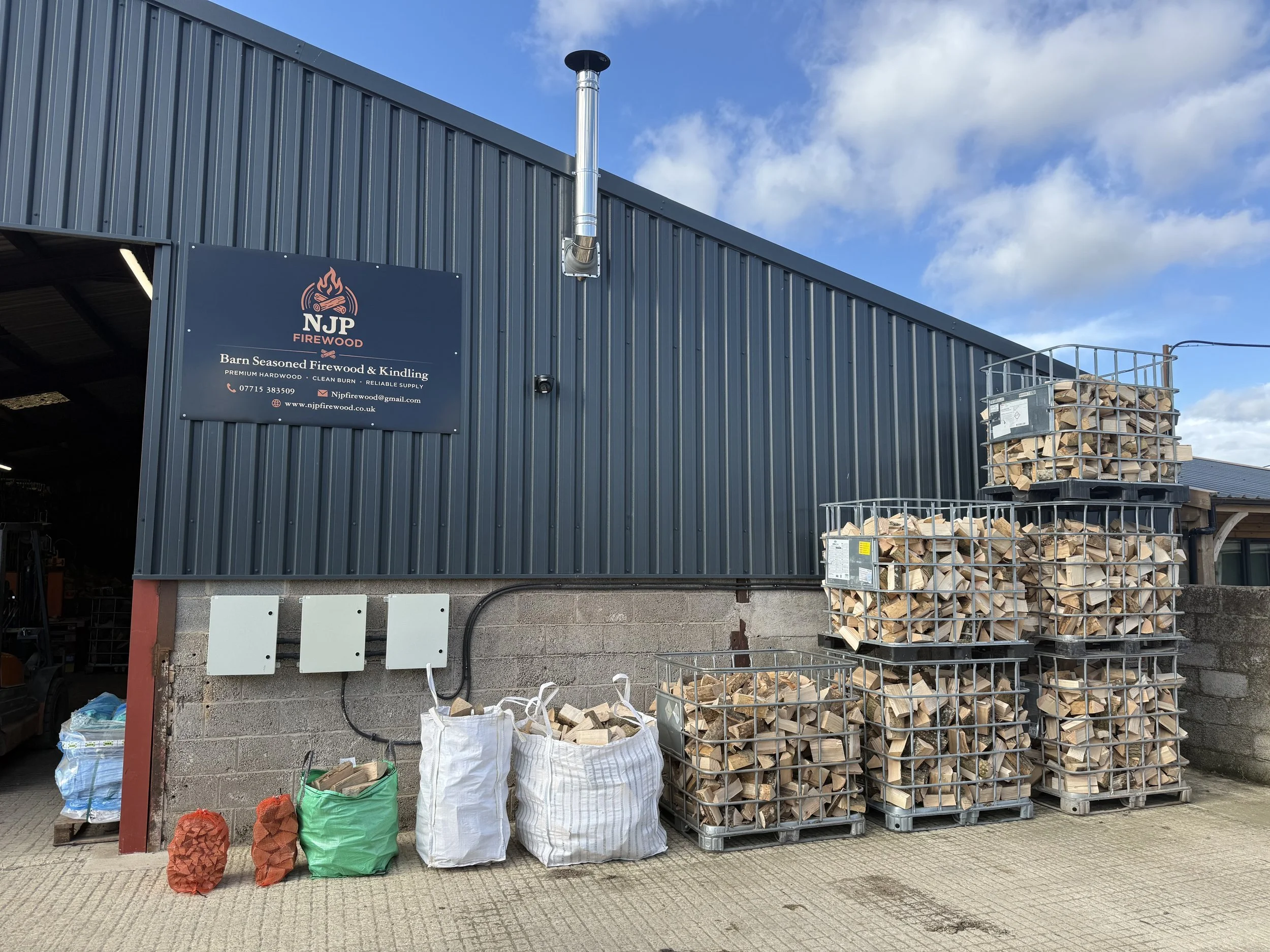 Exterior view of a building with a sign for NJP Firewood, showing stacks of chopped firewood in metal carts and bags of kindling, under a clear sky with some clouds.