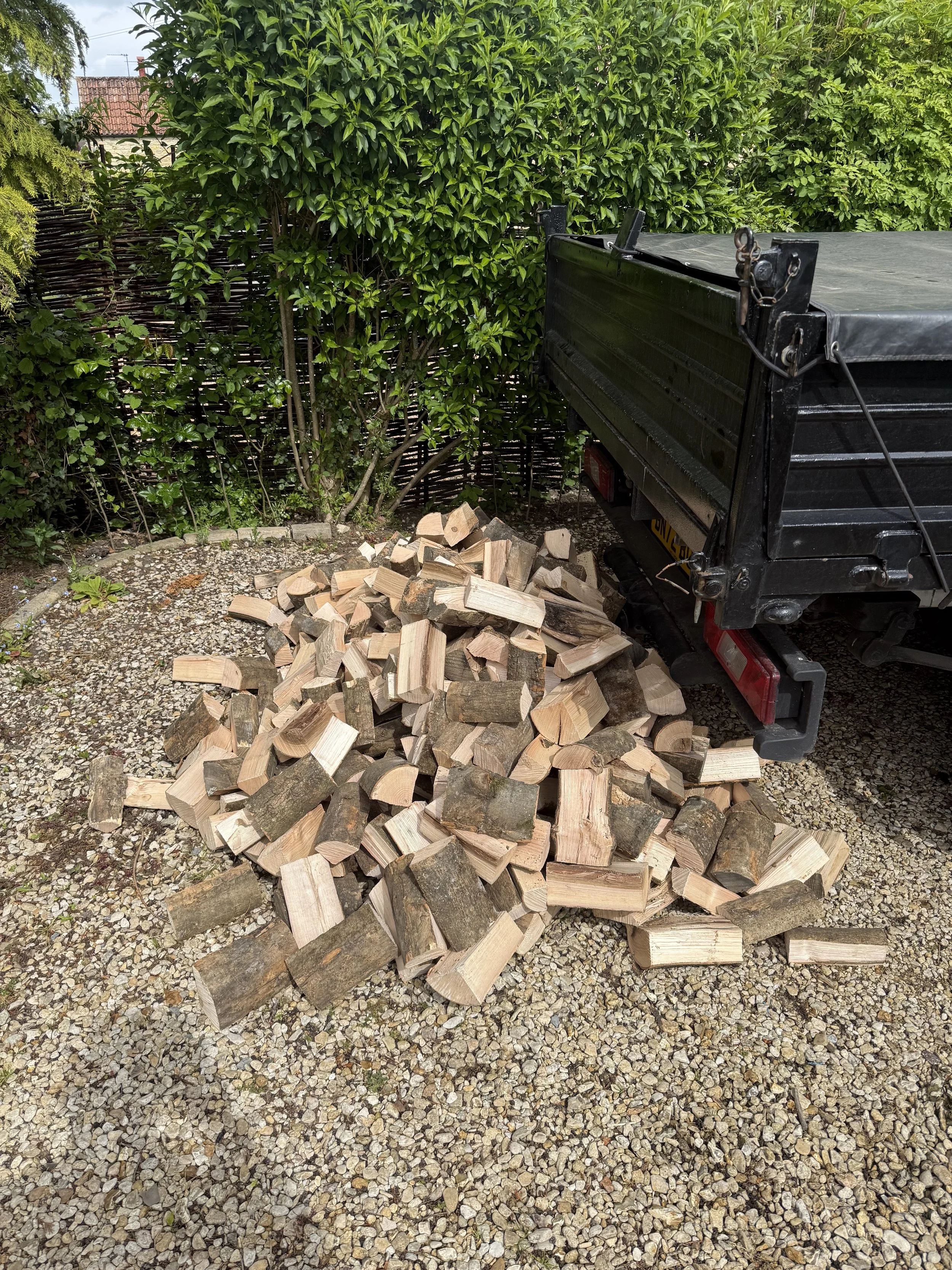A pile of chopped firewood on gravel next to a black truck bed, with bushes and trees in the background.