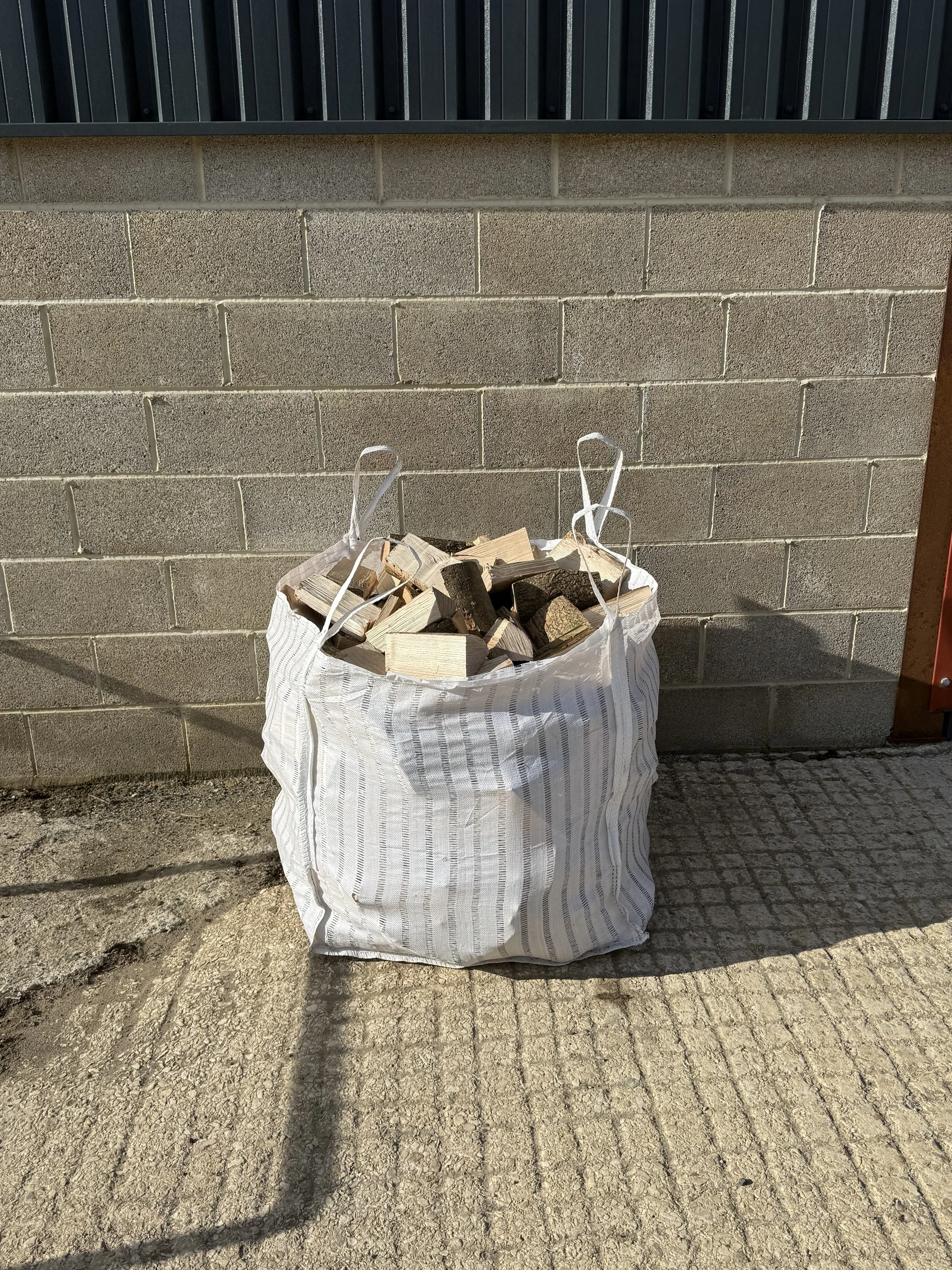 A large white woven bag filled with chopped firewood, placed on a cracked concrete ground next to a brick wall with a black metal fence on top.