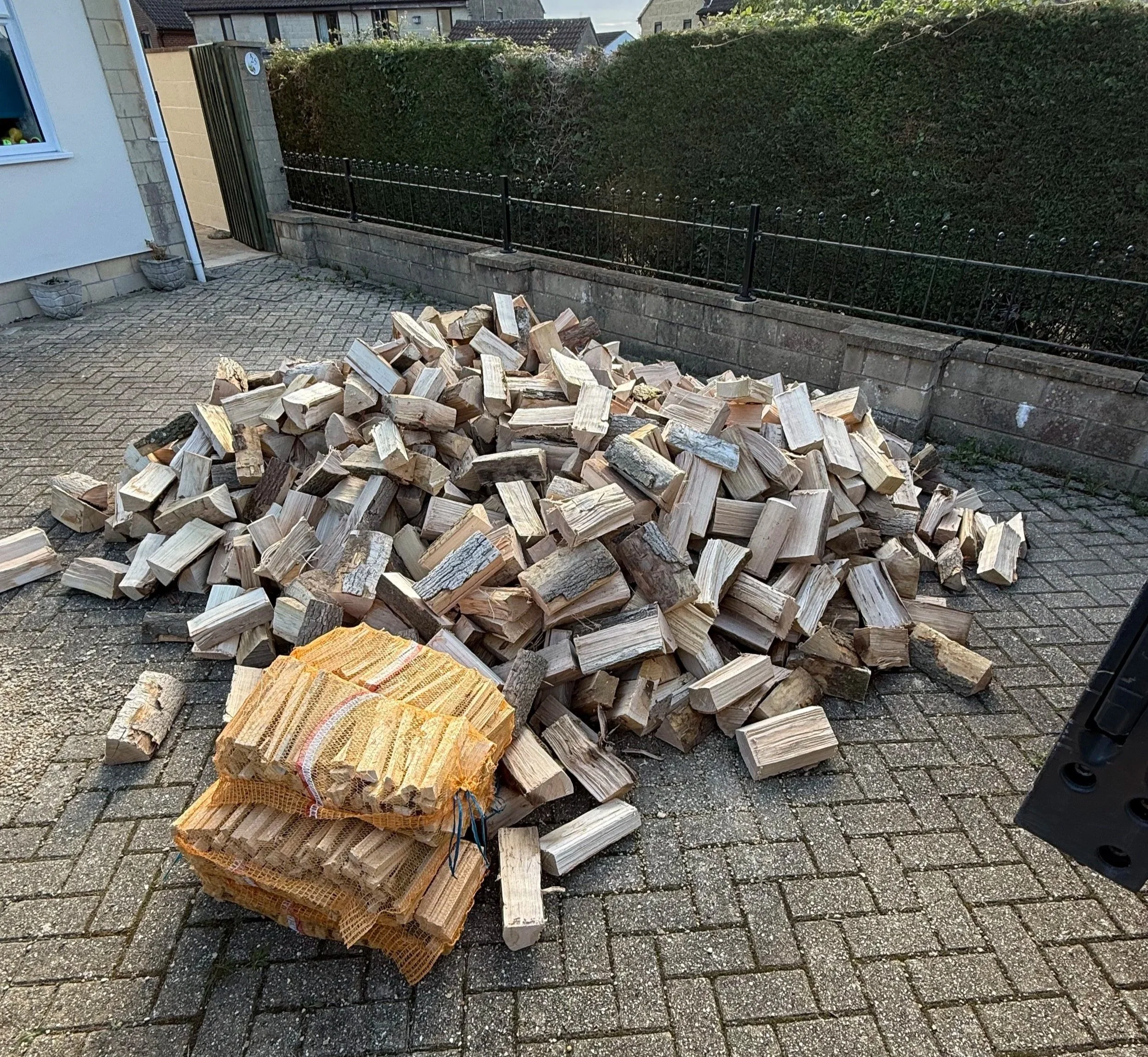 Piles of chopped firewood stacked on a paved outdoor area next to a house with a white wall and window. A burlap sack with additional firewood sits in front of the pile. In the background, there is a black metal fence, green hedge, and neighboring houses.