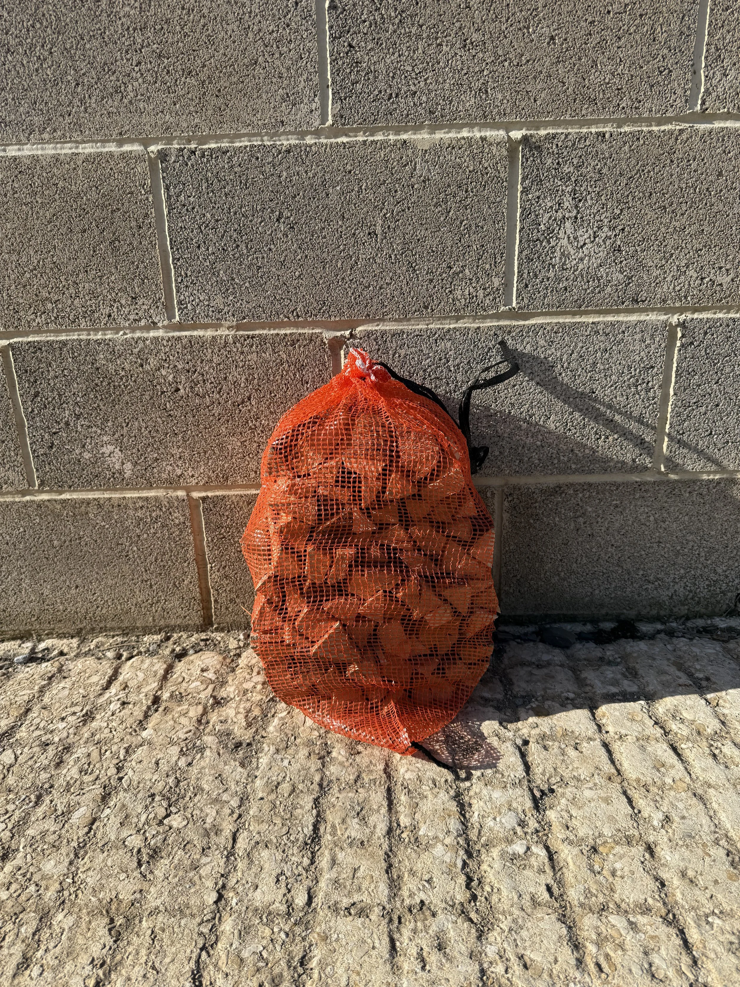 An orange mesh bag filled with trash, hanging against a gray concrete block wall outside on a gravel surface.