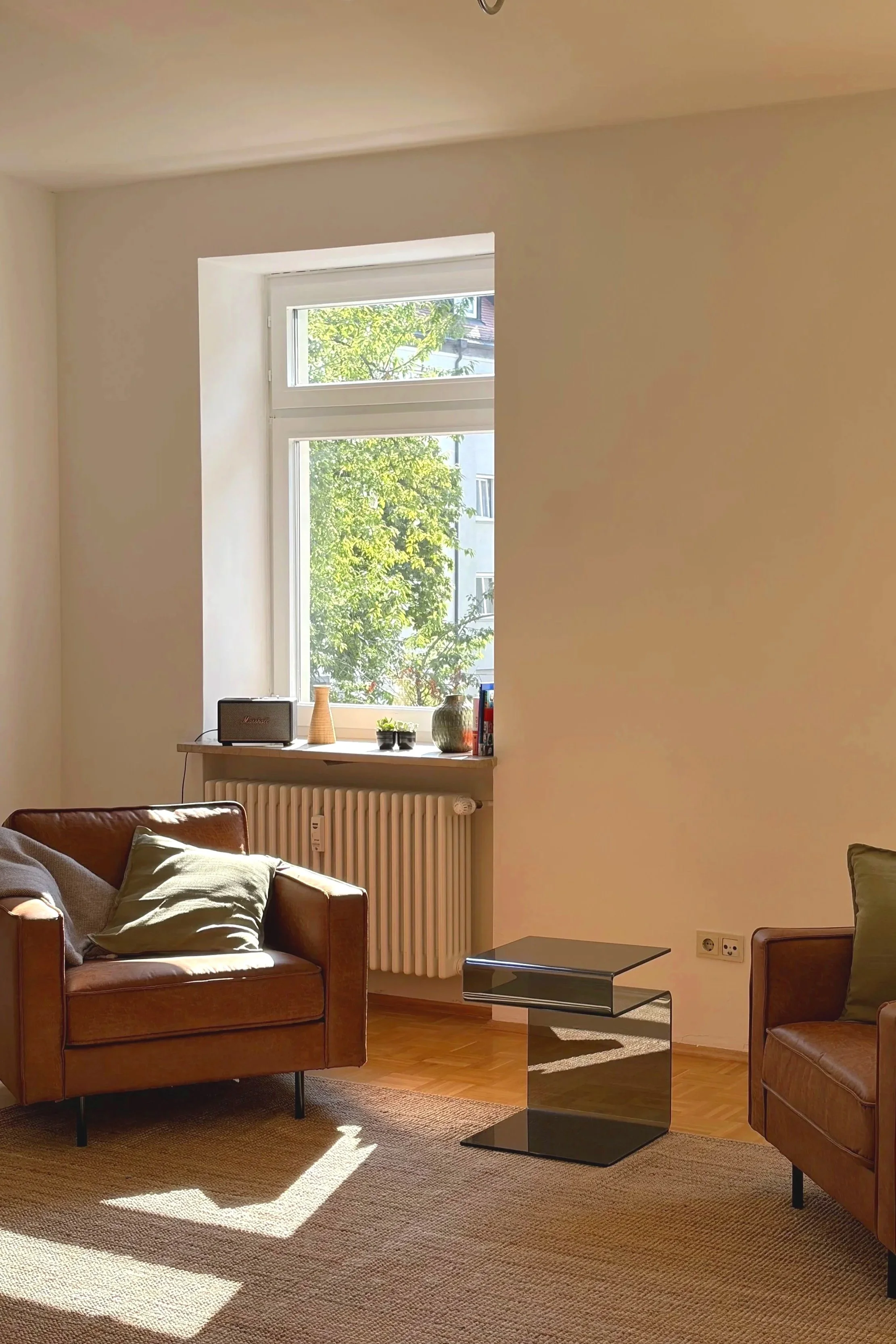 Helles Therapiezimmer mit braunem Sessel, kleinen Kissen, Fenstersims mit dekorativen Gegenständen und Fachliteratur, Jute Teppich, kleinem Glas Tisch und Fenster mit Blick auf grüne Bäume.