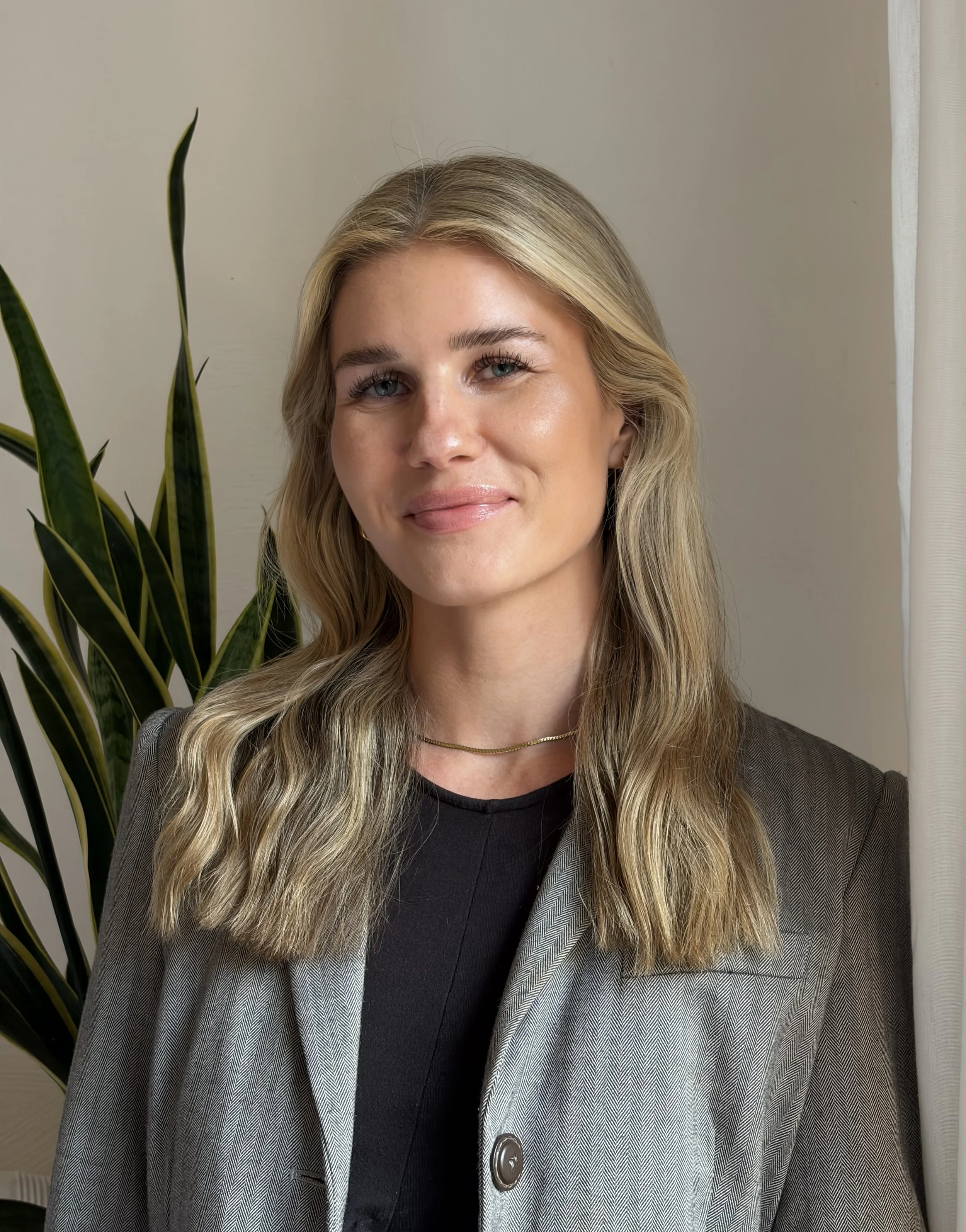 Junge blonde Psychotherapeutin mit einem schwarzen Oberteil und einem grauen Blazer, steht vor weißem Hintergrund neben einer Grünpflanze.