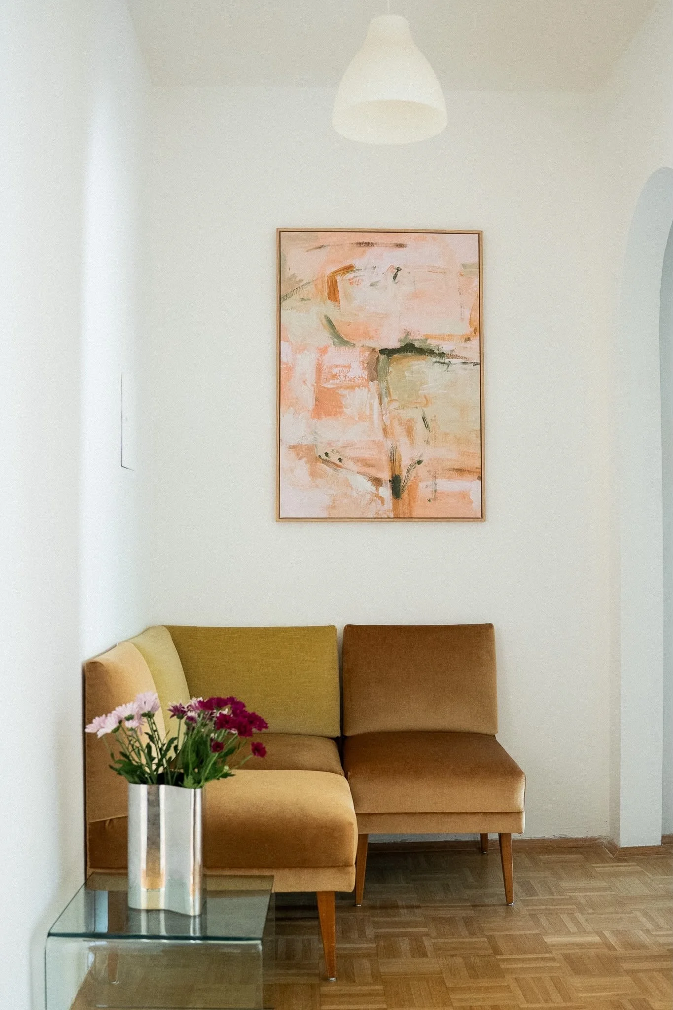 Ein Wohnraum mit einer beigen und braunen Couch, acker und einem abstrakten Bild an der Wand, eine Vase mit bunten Blumen auf einem kleinen Glastisch.