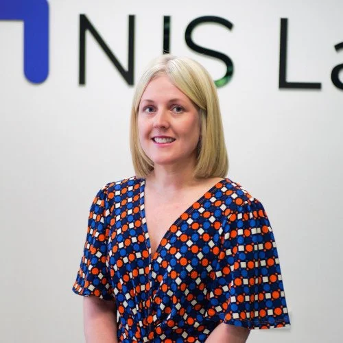 A woman with shoulder-length blonde hair, wearing a colorful geometric patterned dress, standing in front of a white wall with partially visible black text.