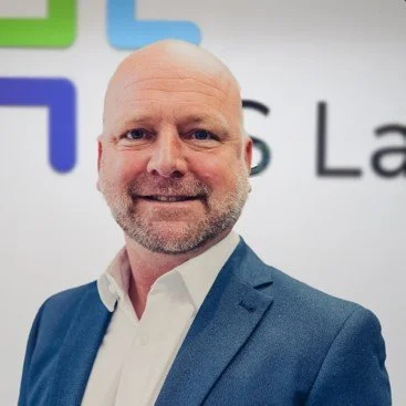 A bald man with a beard, wearing a blue suit and white shirt, smiling in front of a blurred background with a partial logo.