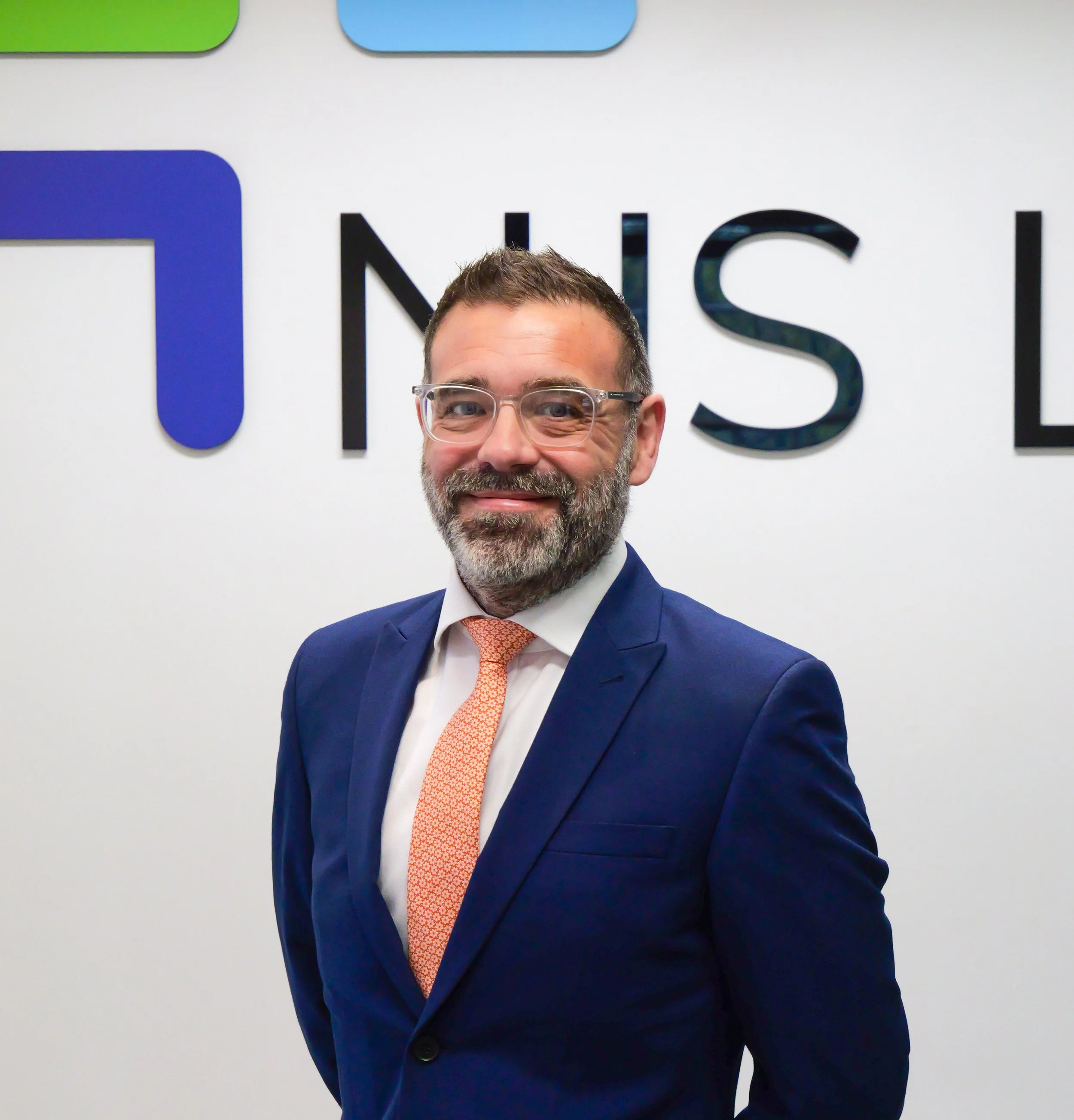 Man wearing glasses, a navy blue suit, white shirt, and orange patterned tie, smiling, standing in front of a white wall with colorful logo elements and black text.