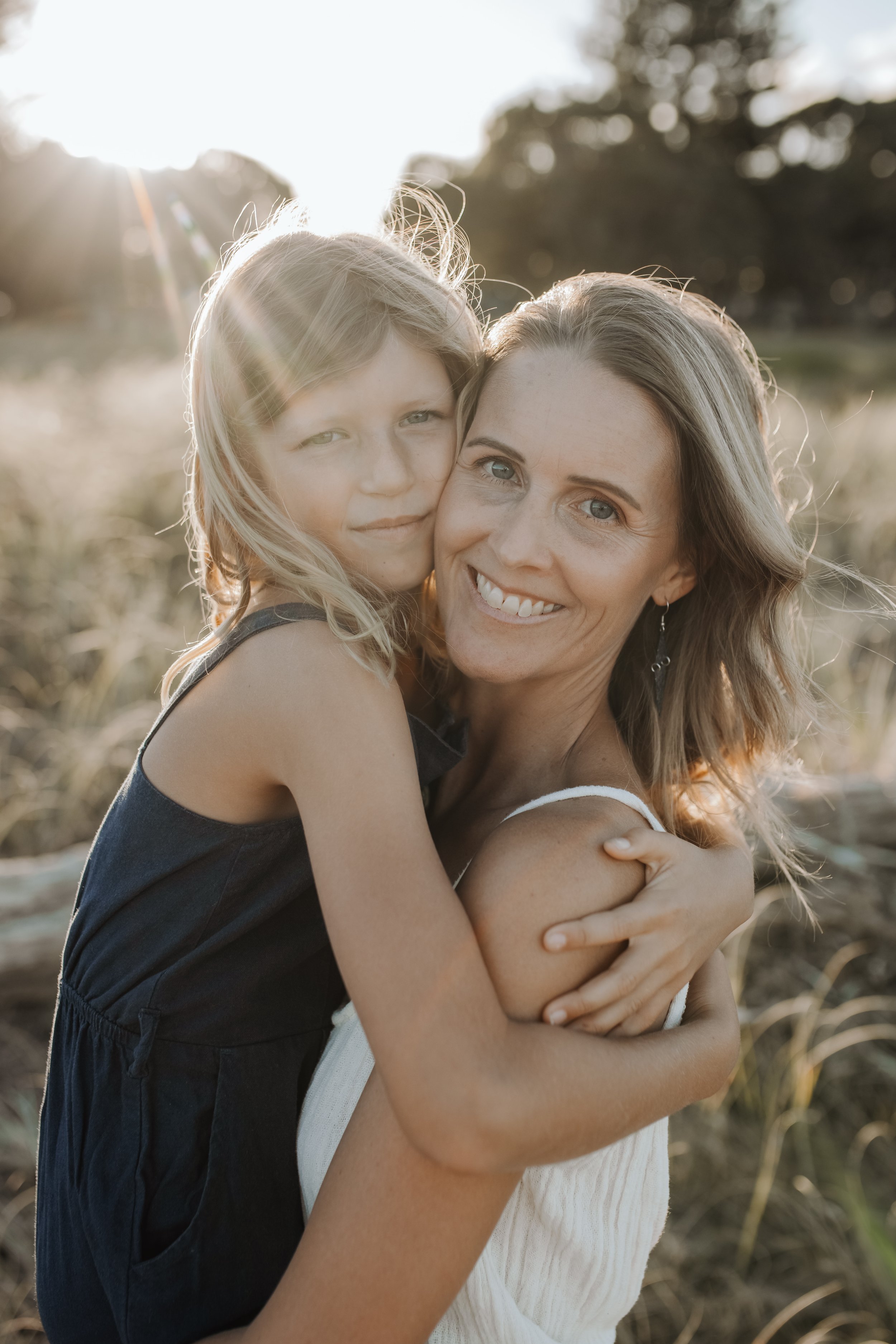 A woman and a young girl are embracing each other outdoors during sunset, with sunlight shining from behind them and trees in the background.