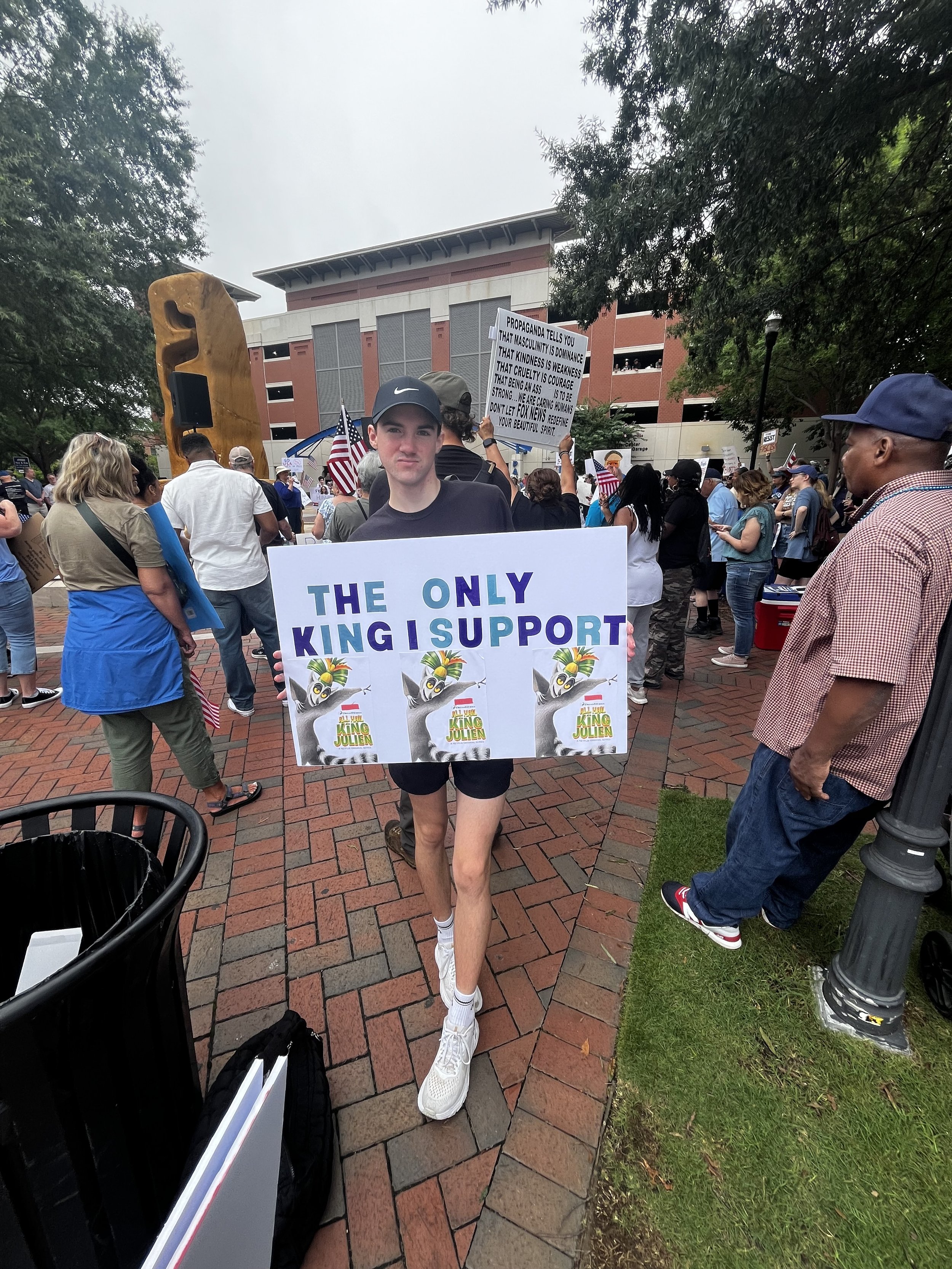 A young person wearing a black Nike cap and white sneakers holding a sign that reads, 'The only king I support,' with colorful wording and cartoon animal illustrations, during a protest or rally with a crowd holding signs and flags in an outdoor urban setting with brick ground and trees.