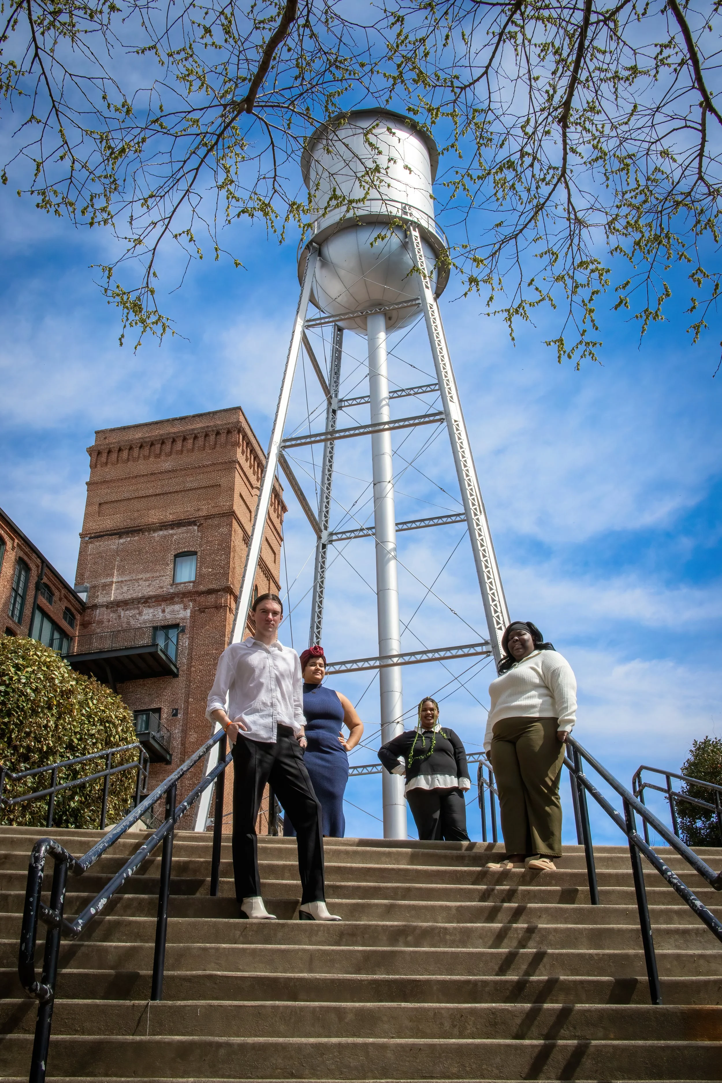 Sam and the Muscogee County Young Democrats Board standing on outdoor steps, with a large water tower, blue sky, a brick building and trees in the background.