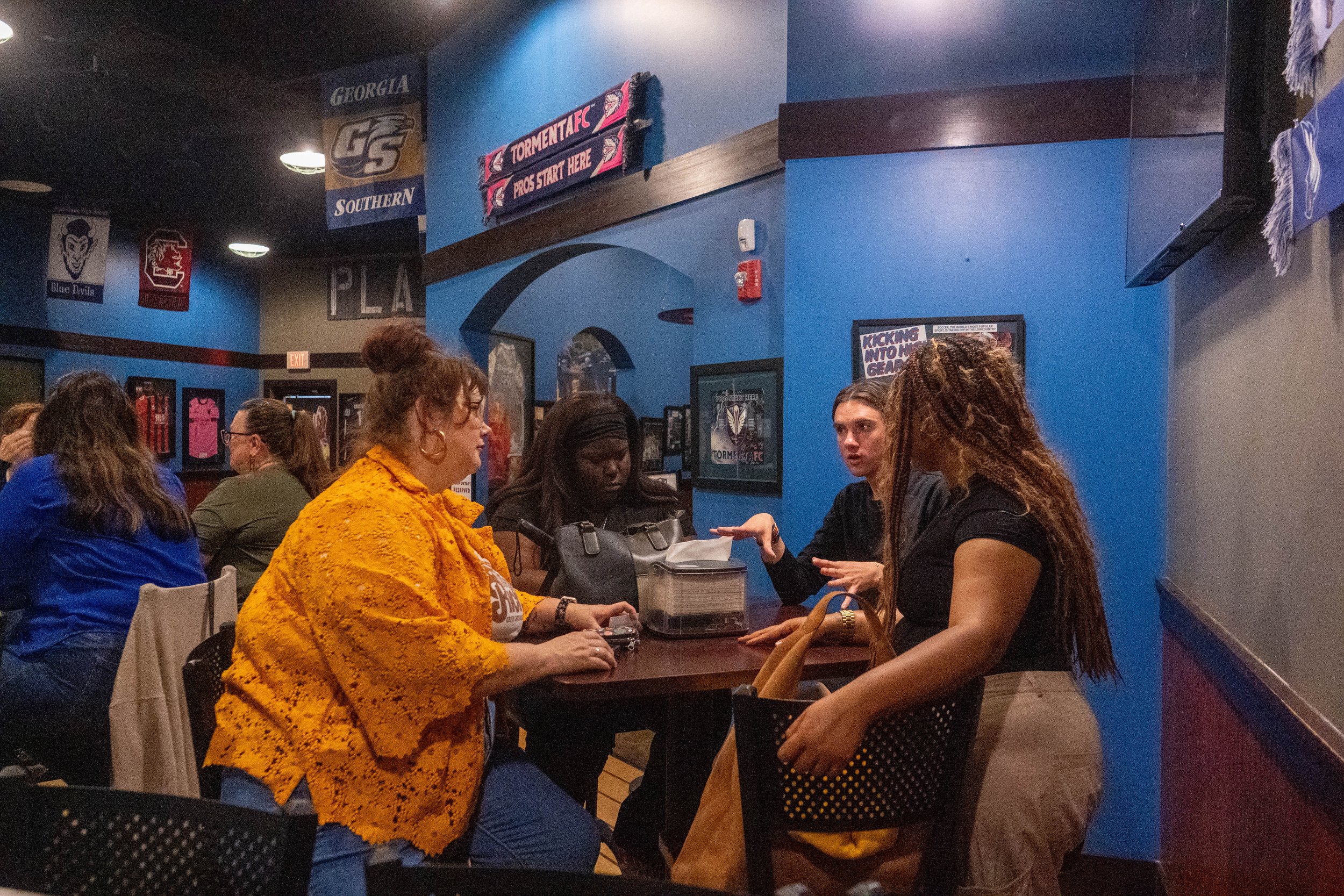 A group of women sitting at a table inside a sports-themed restaurant or bar, with blue walls adorned with sports team banners and posters.