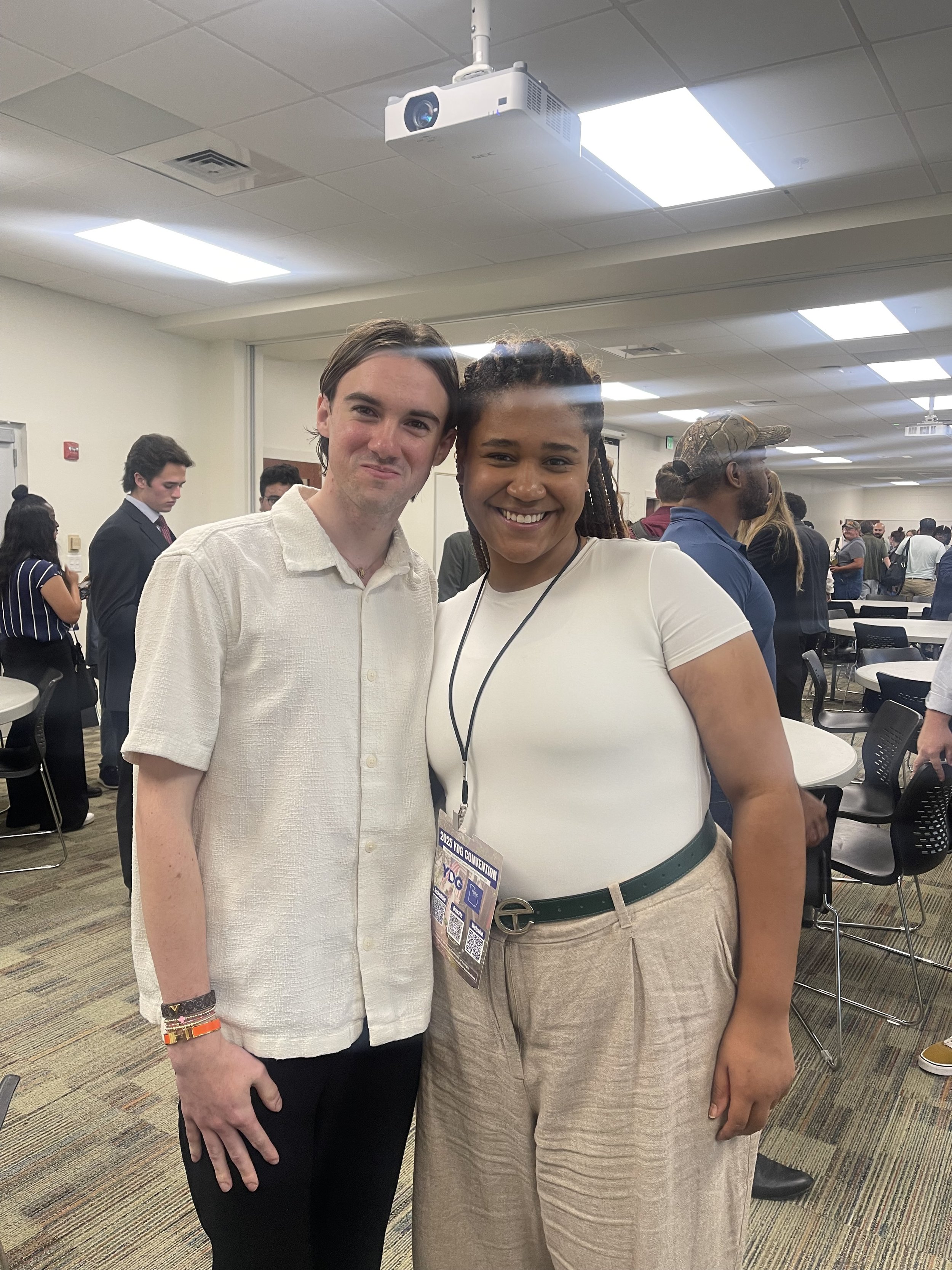Two people, a man and a woman, are smiling and standing together at a conference or event in a room with many other attendees. The man has dark hair and is wearing a white shirt, while the woman has braids and is wearing a white top with beige pants and a green belt. The woman has a conference badge hanging from her neck.